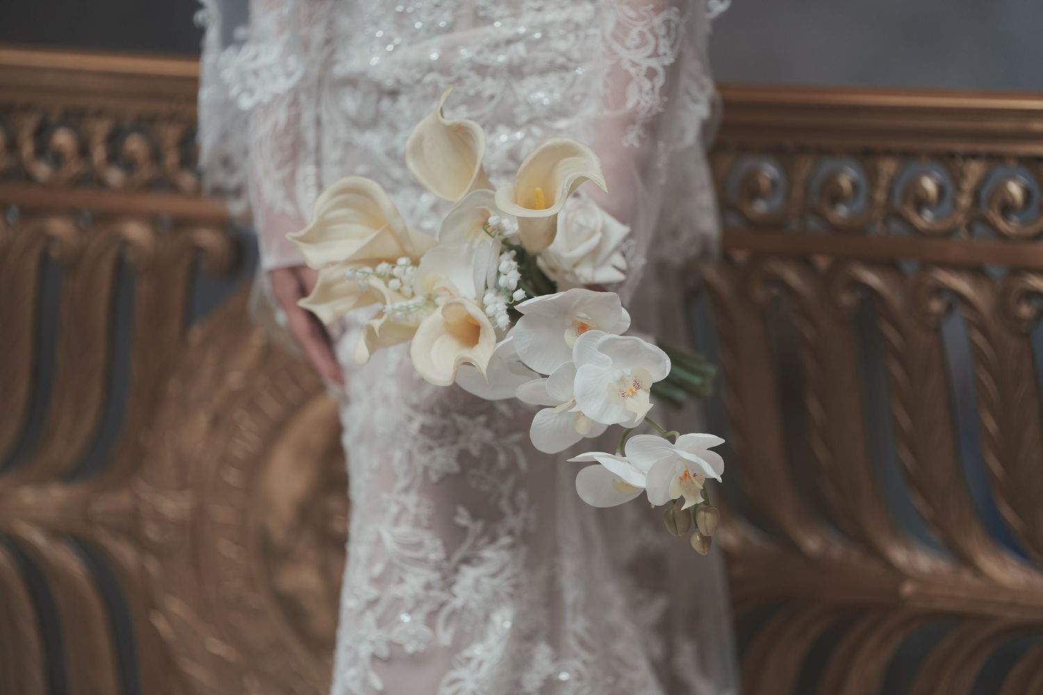 Bride holding elegant white calla lily and orchid bouquet against lace wedding gown near ornate wooden decor.