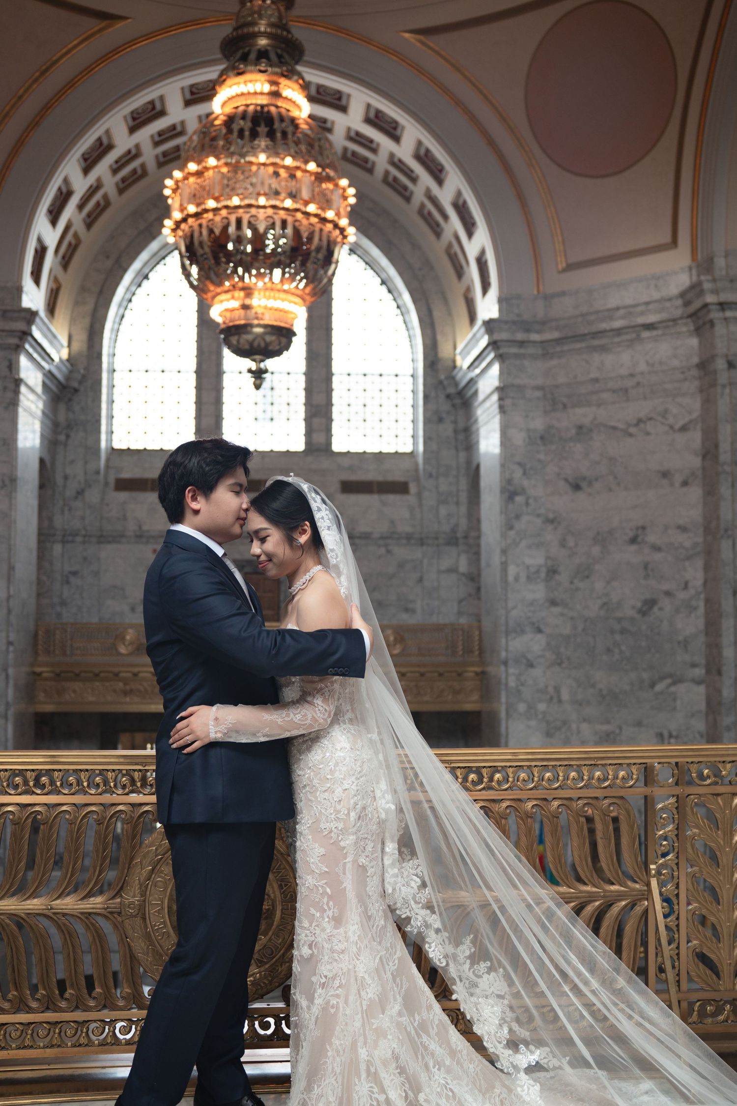 Bride and groom embracing under ornate chandelier on grand staircase in elegant marble wedding venue.