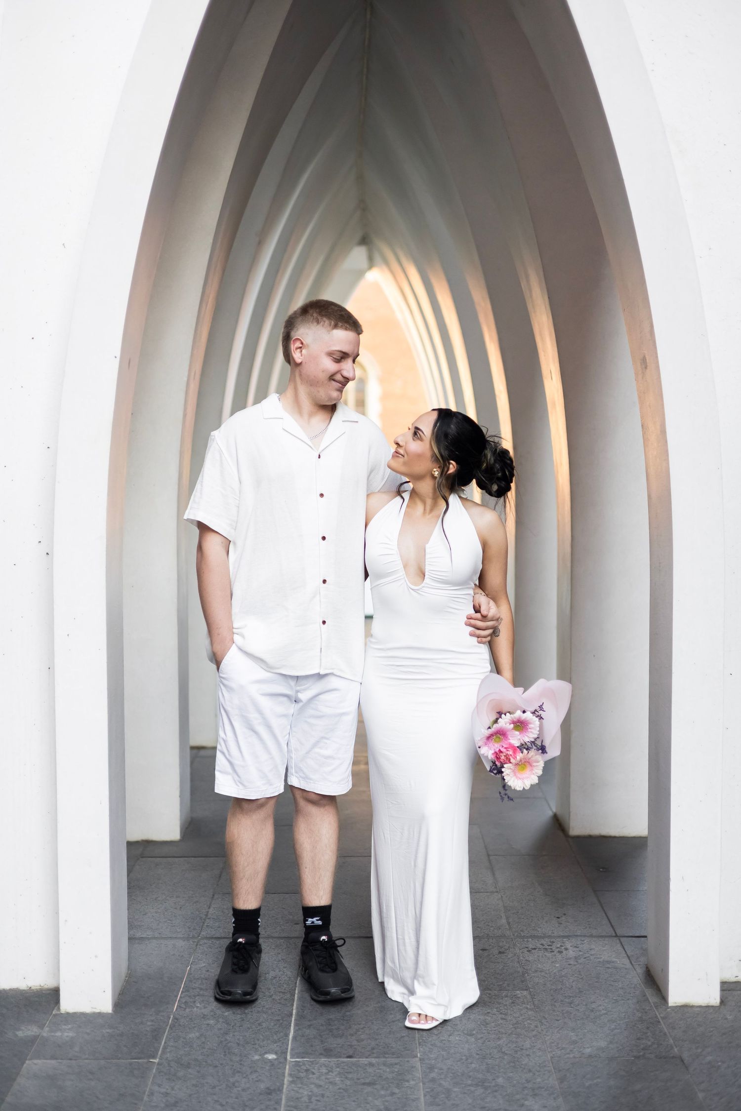 Couple in white outfits posing under elegant arched corridor, woman holding pink bouquet, sharing a romantic moment.