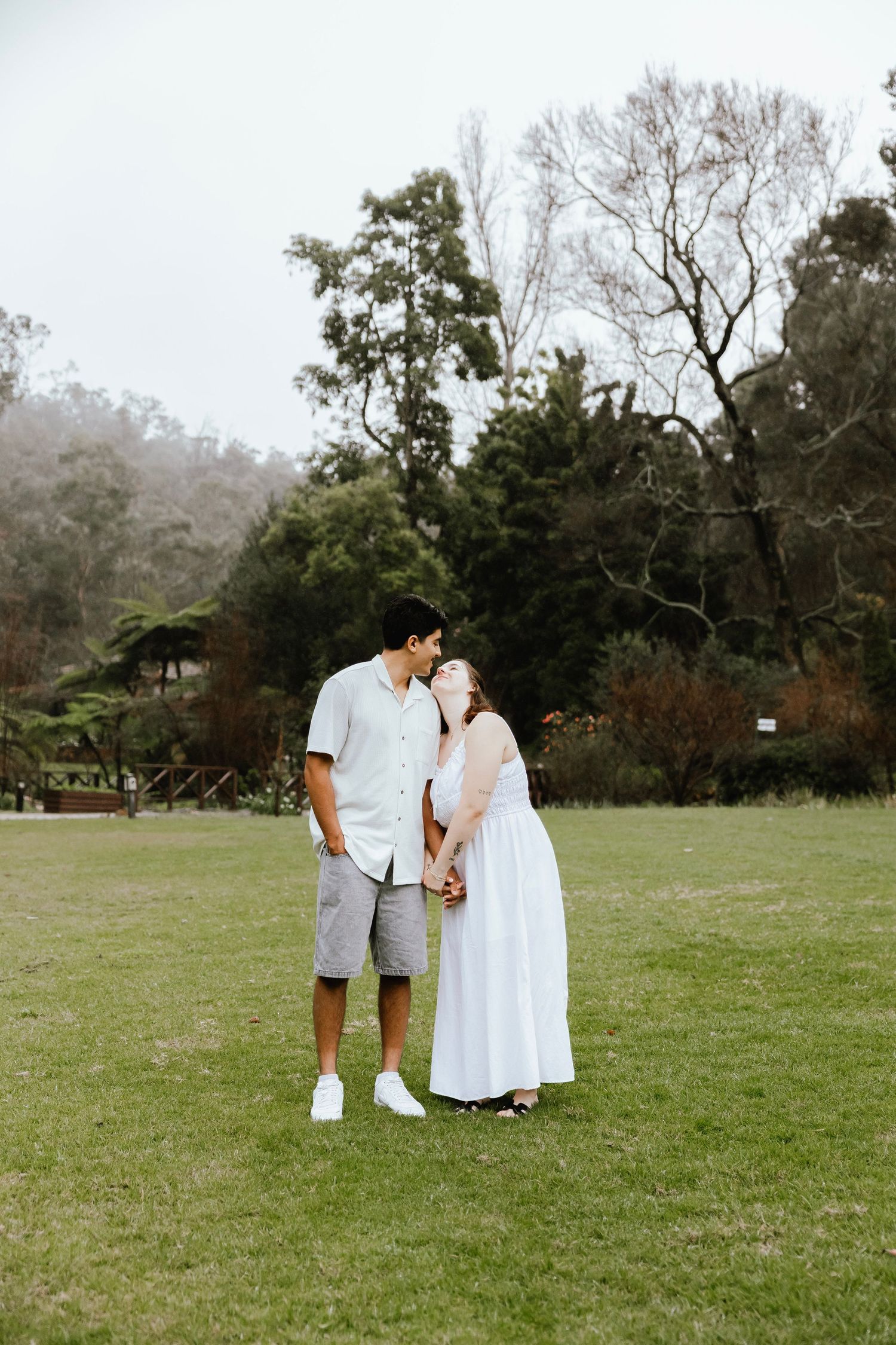Couple sharing a tender kiss while holding hands on a green lawn in a misty park.