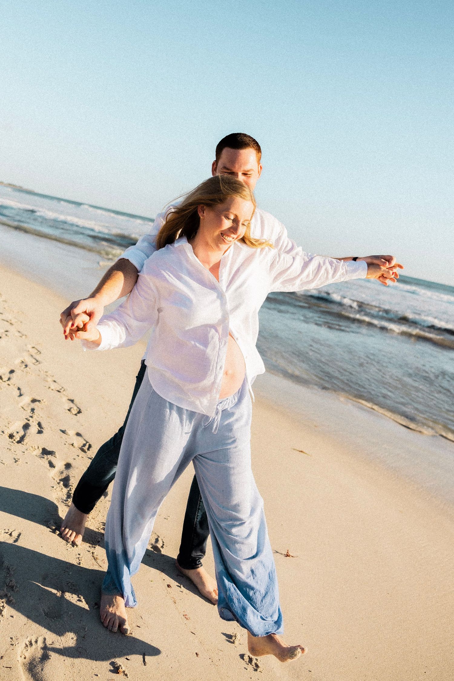 Joyful pregnant couple dancing playfully with arms outstretched on a sunny sandy beach.