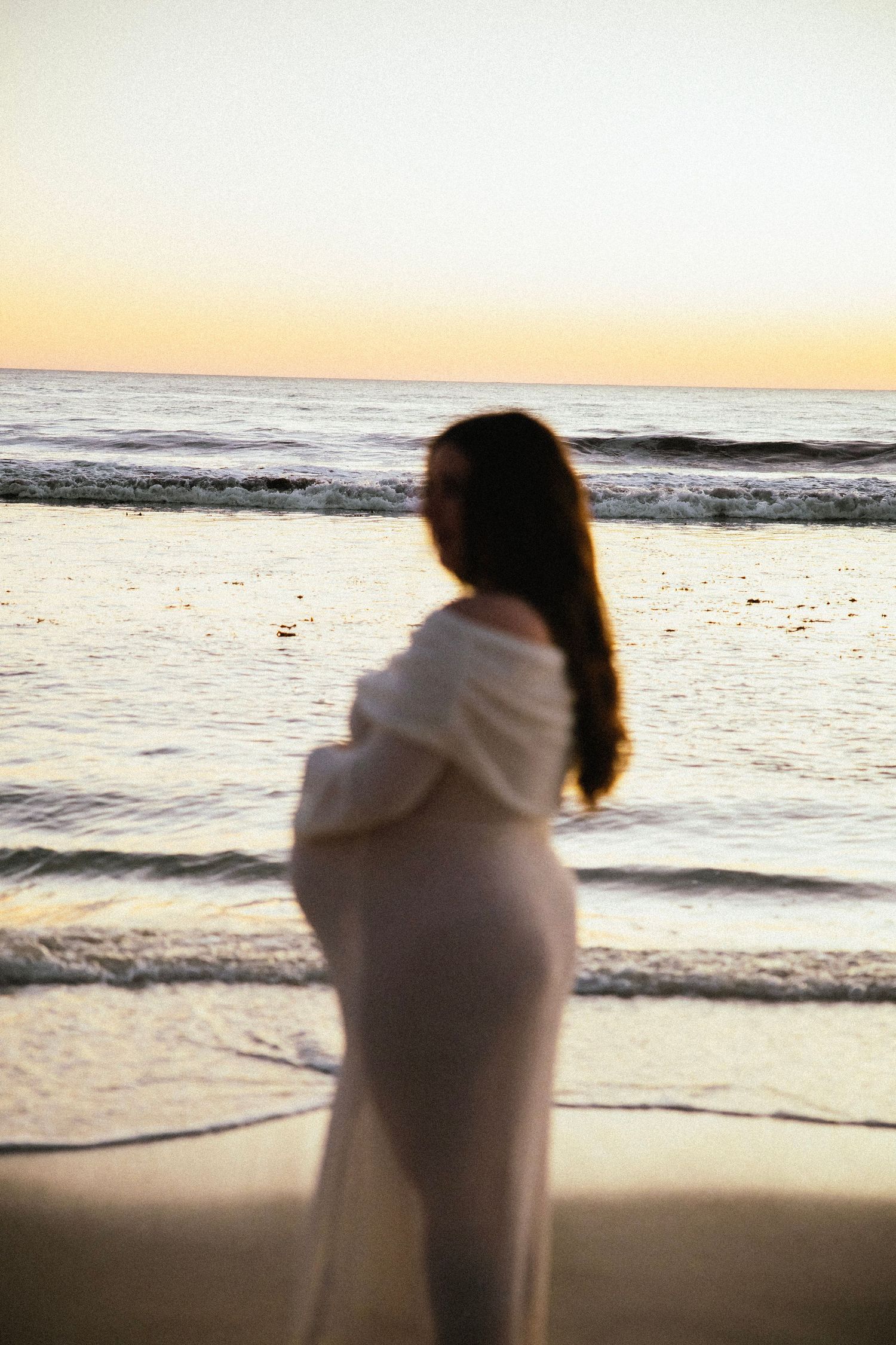 Pregnant woman in white dress standing at ocean shore during golden sunset, silhouetted against calm waves.