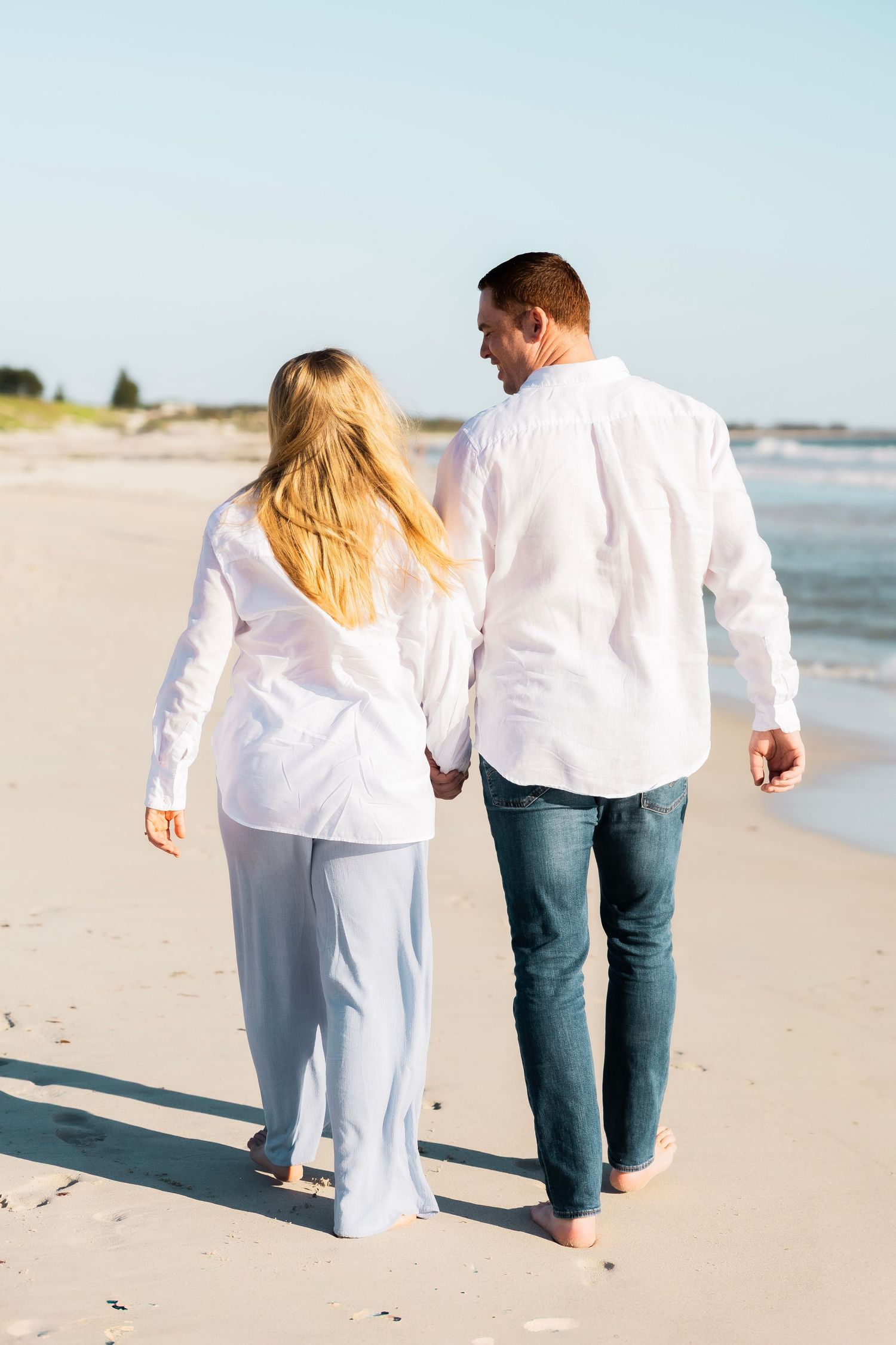 Couple in white shirts holding hands walking barefoot on a sunny beach, viewed from behind.