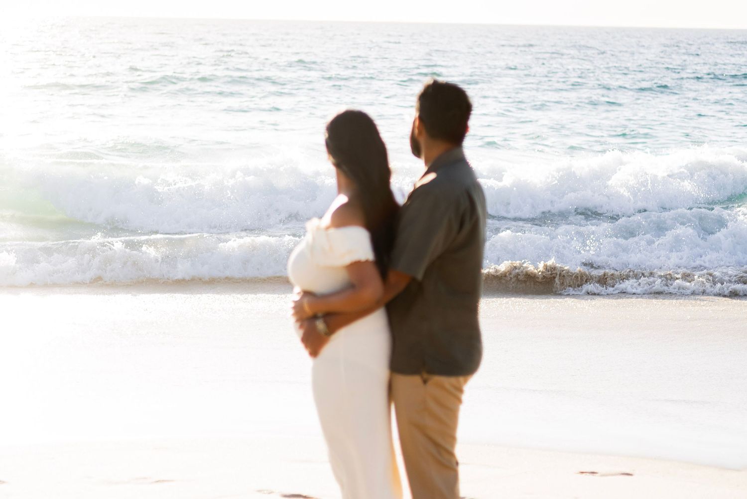 Couple embracing on a sunny beach, woman in white maternity dress, waves crashing behind them.