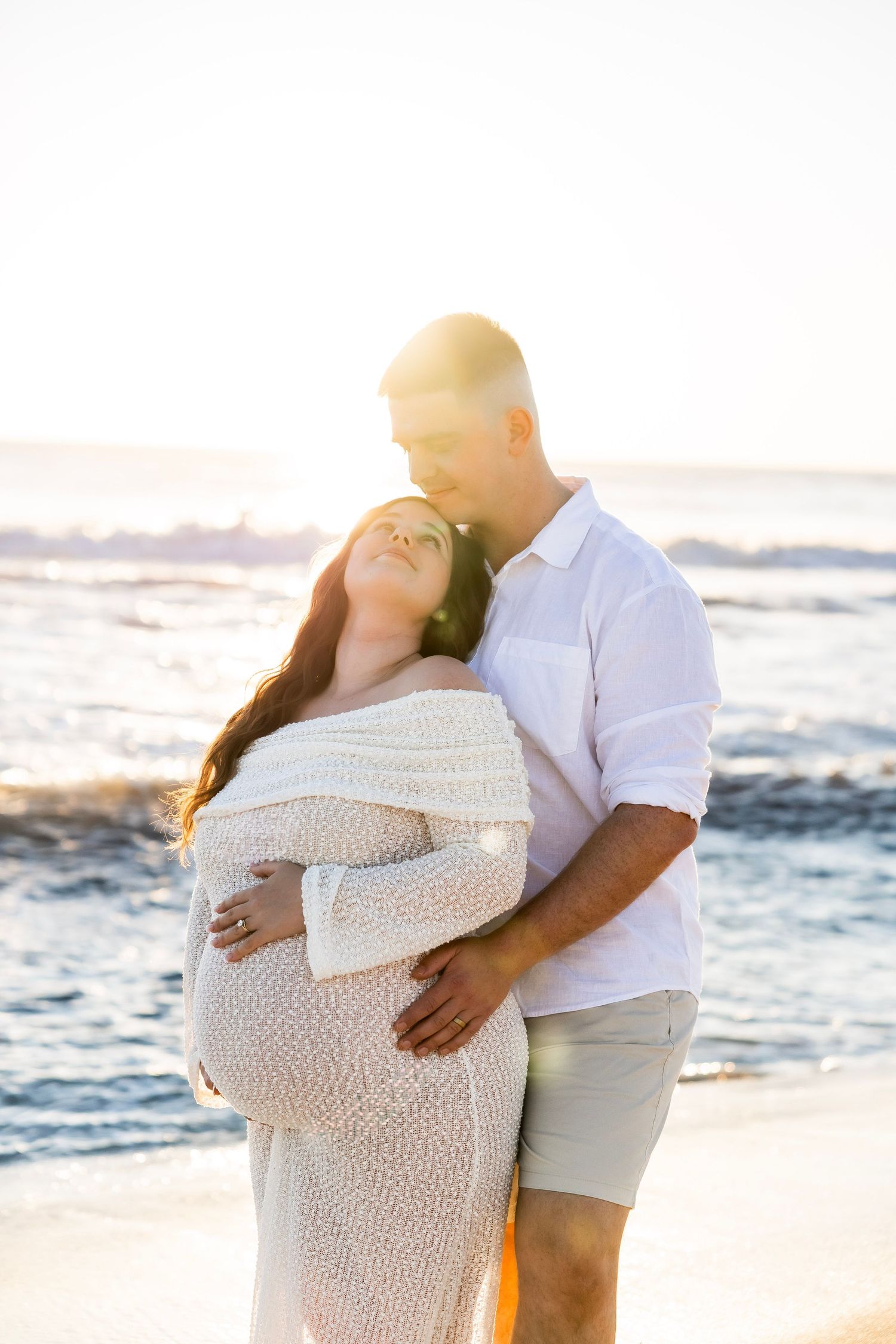 Pregnant couple embracing on beach at golden sunset, woman in white knit dress leaning back against man in white shirt.