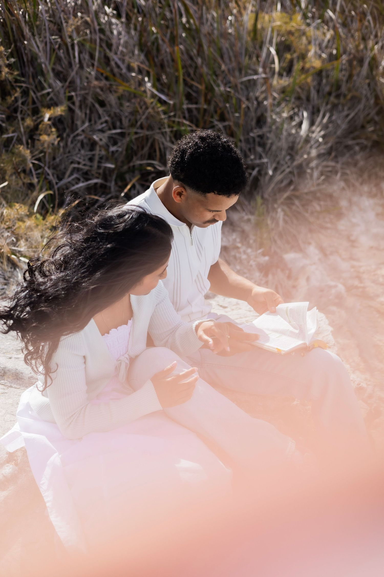 Couple dressed in white sitting together on sandy ground surrounded by tall grass in bright sunlight.
