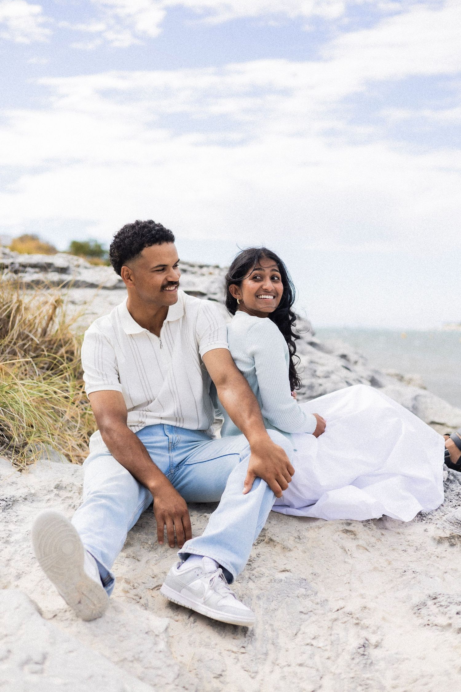 A happy couple sits together on sandy rocks by the ocean, smiling and enjoying the view.