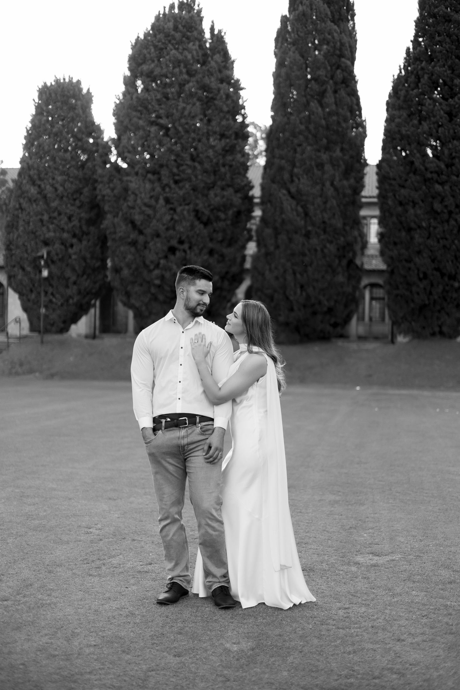 Black and white photo of couple standing on lawn, woman in white dress touching mans face near cypress trees.