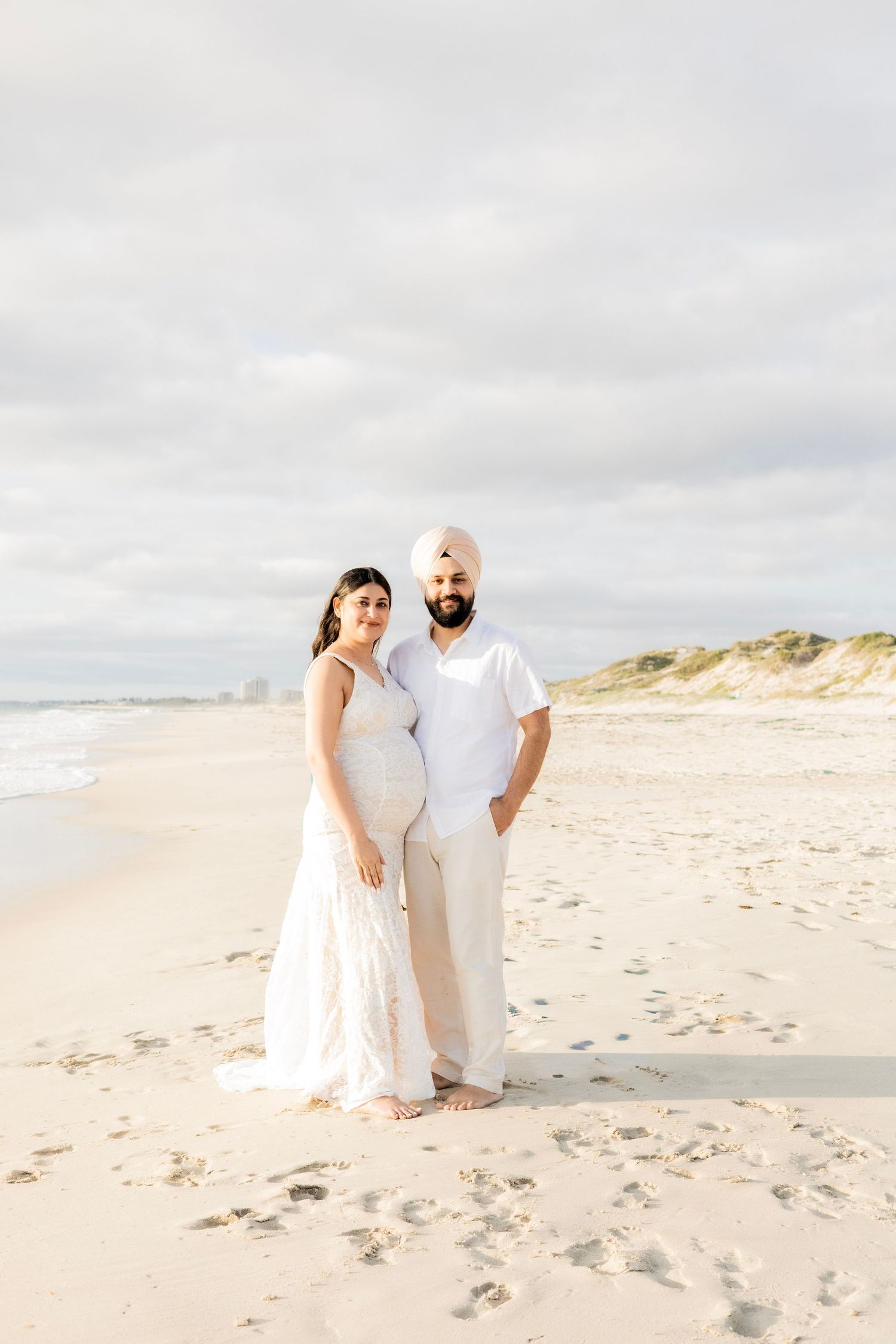 Expecting couple standing on sandy beach, man in turban and white shirt beside pregnant woman in white dress.