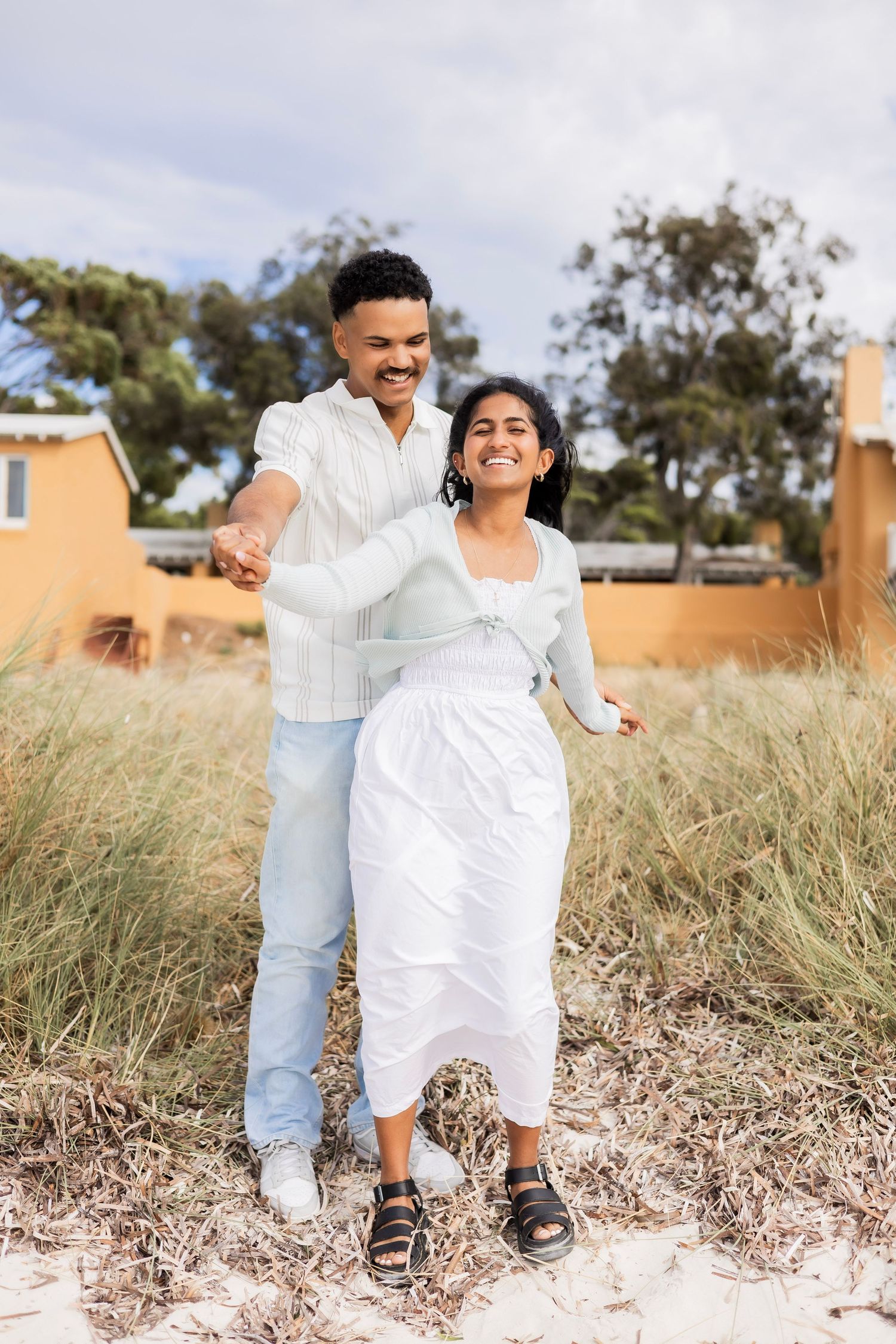 Happy couple holding hands and dancing on sandy beach grass with orange building backdrop.