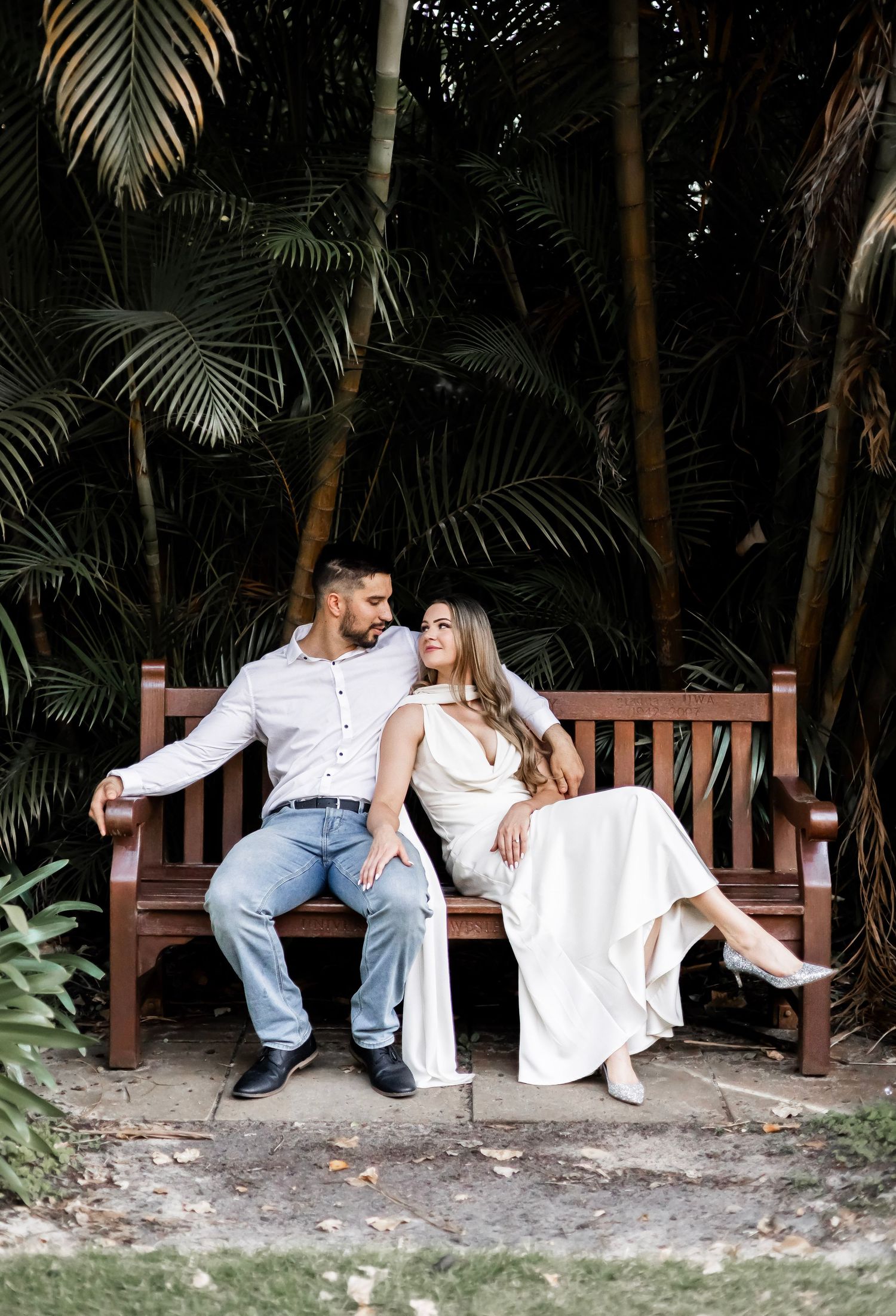 Romantic couple sitting together on a wooden bench surrounded by lush tropical palm trees and foliage.