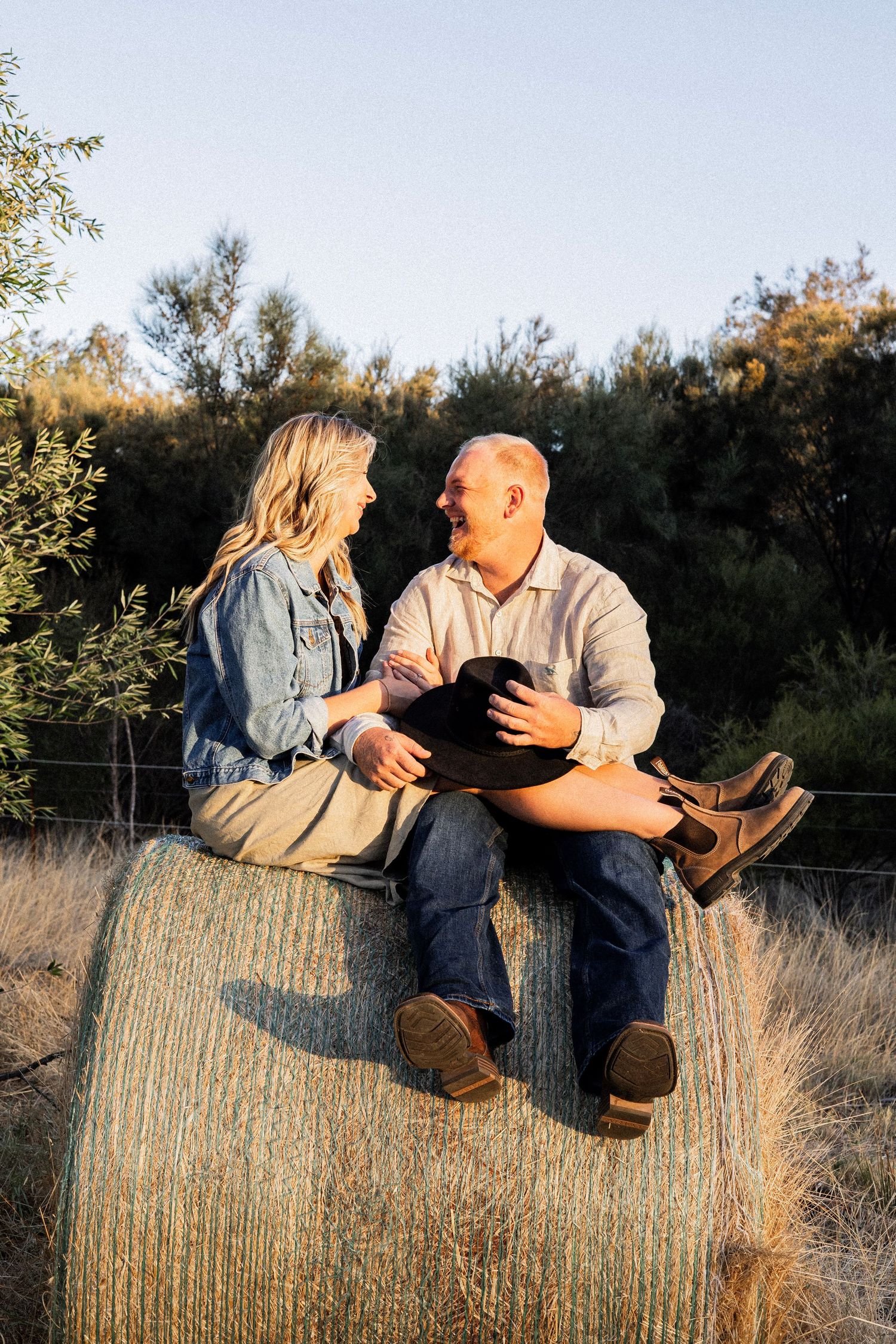 Couple laughing together while sitting on a hay bale in a rural field at golden hour.