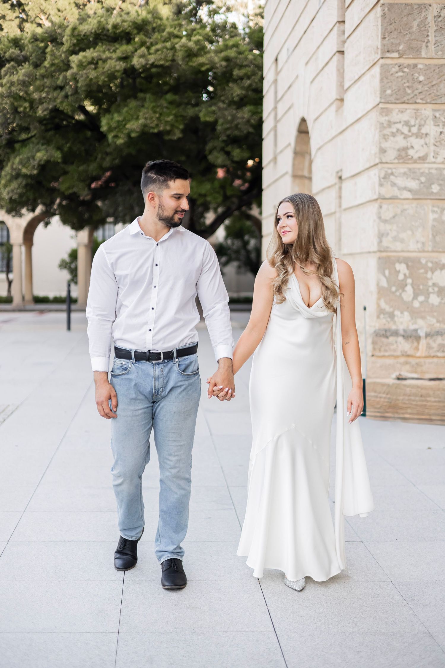 Engaged couple holding hands walking together, man in white shirt and jeans, woman in white satin gown.