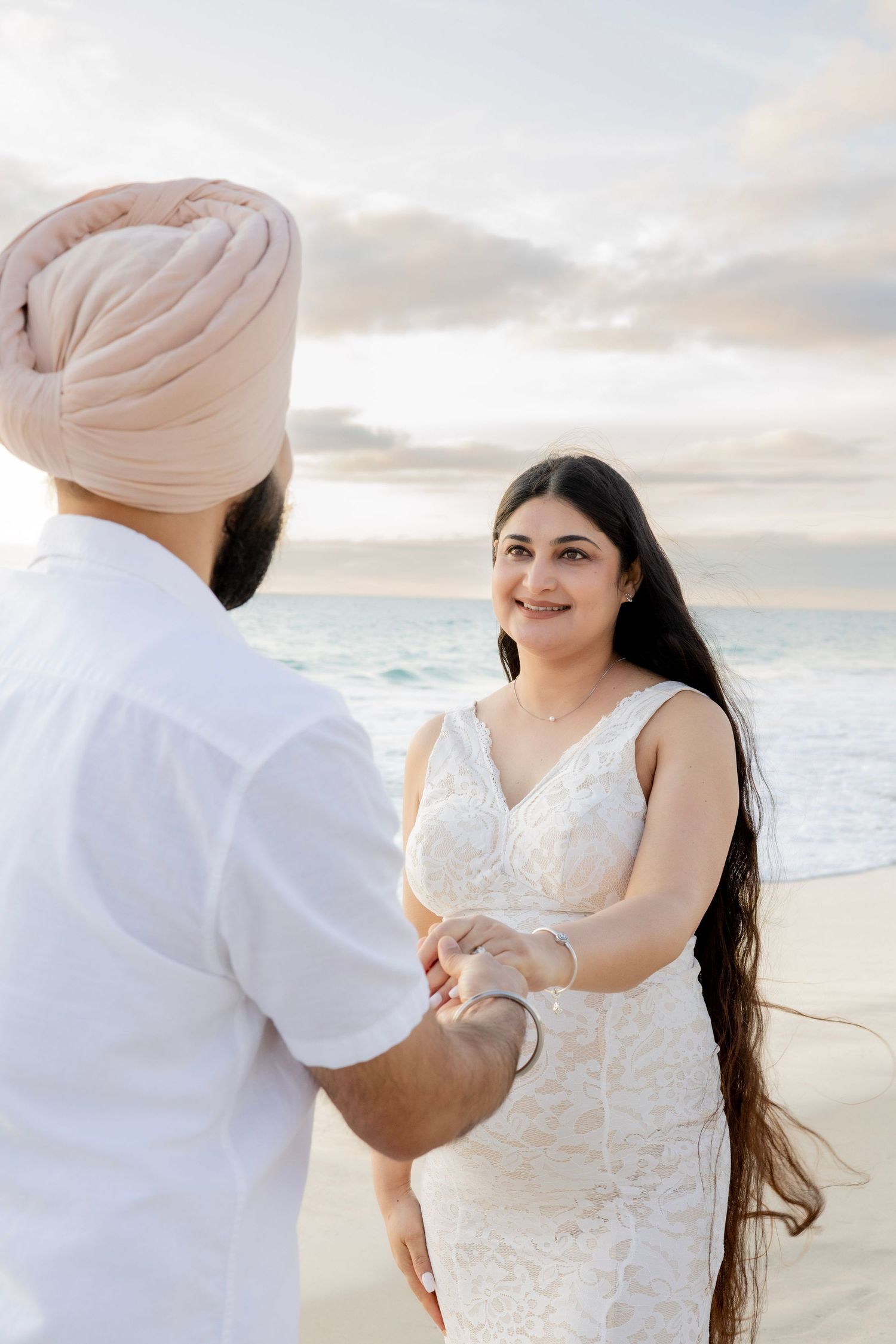 Pregnant woman in white lace dress smiling at her partner on a beach at sunset, holding hands.