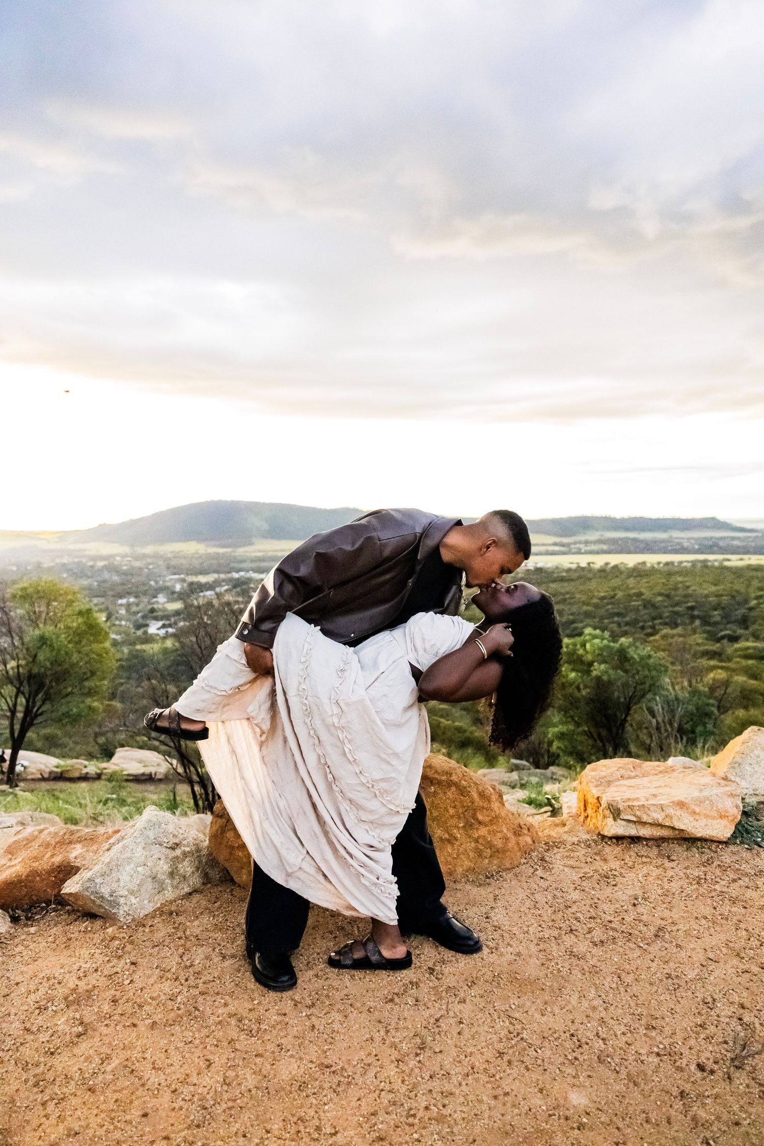 Couple sharing a romantic dip kiss on a rocky hilltop overlook with scenic valley and mountains at sunset.