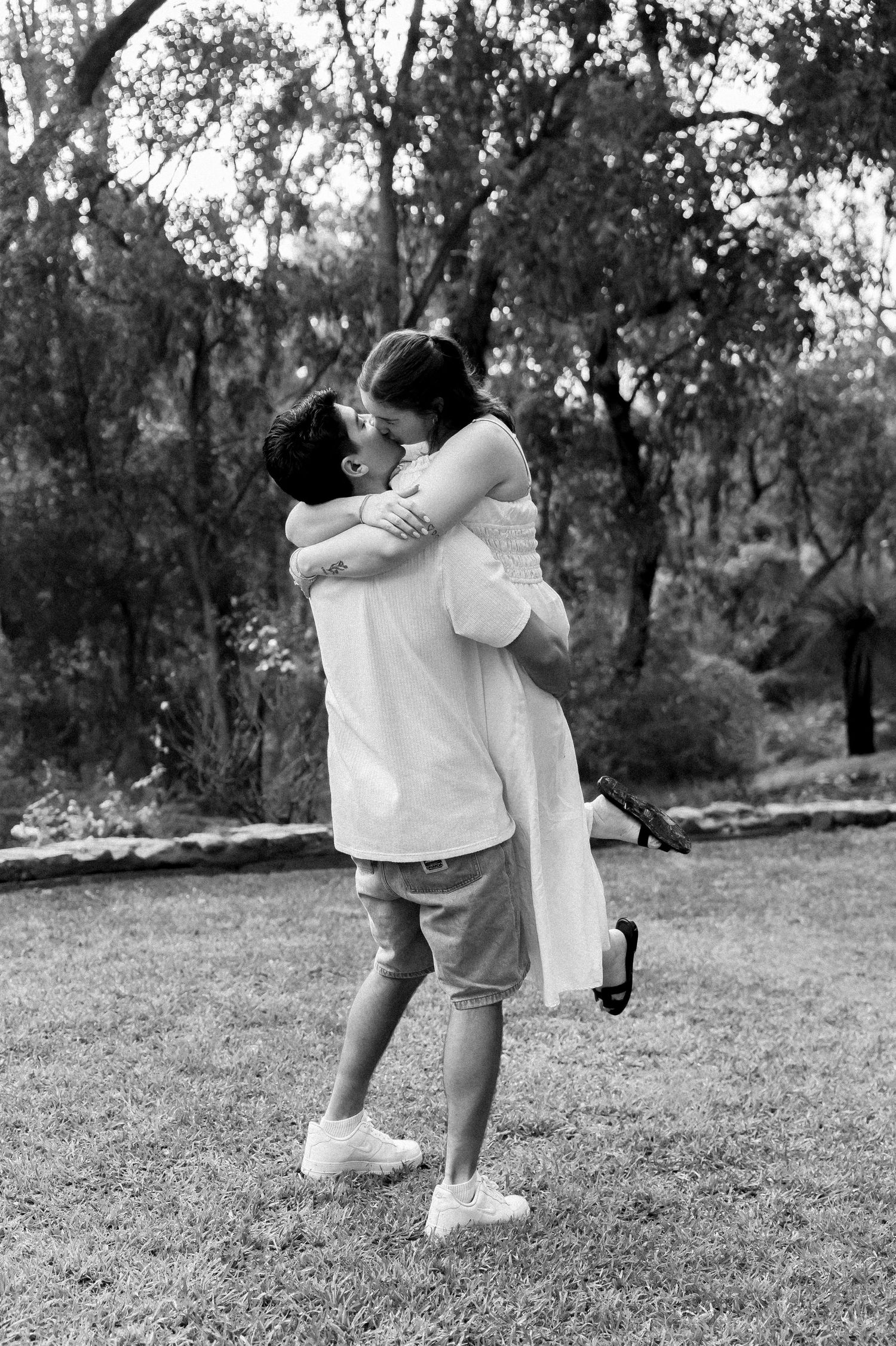 Black and white photo of couple embracing and kissing as man lifts woman in outdoor park.