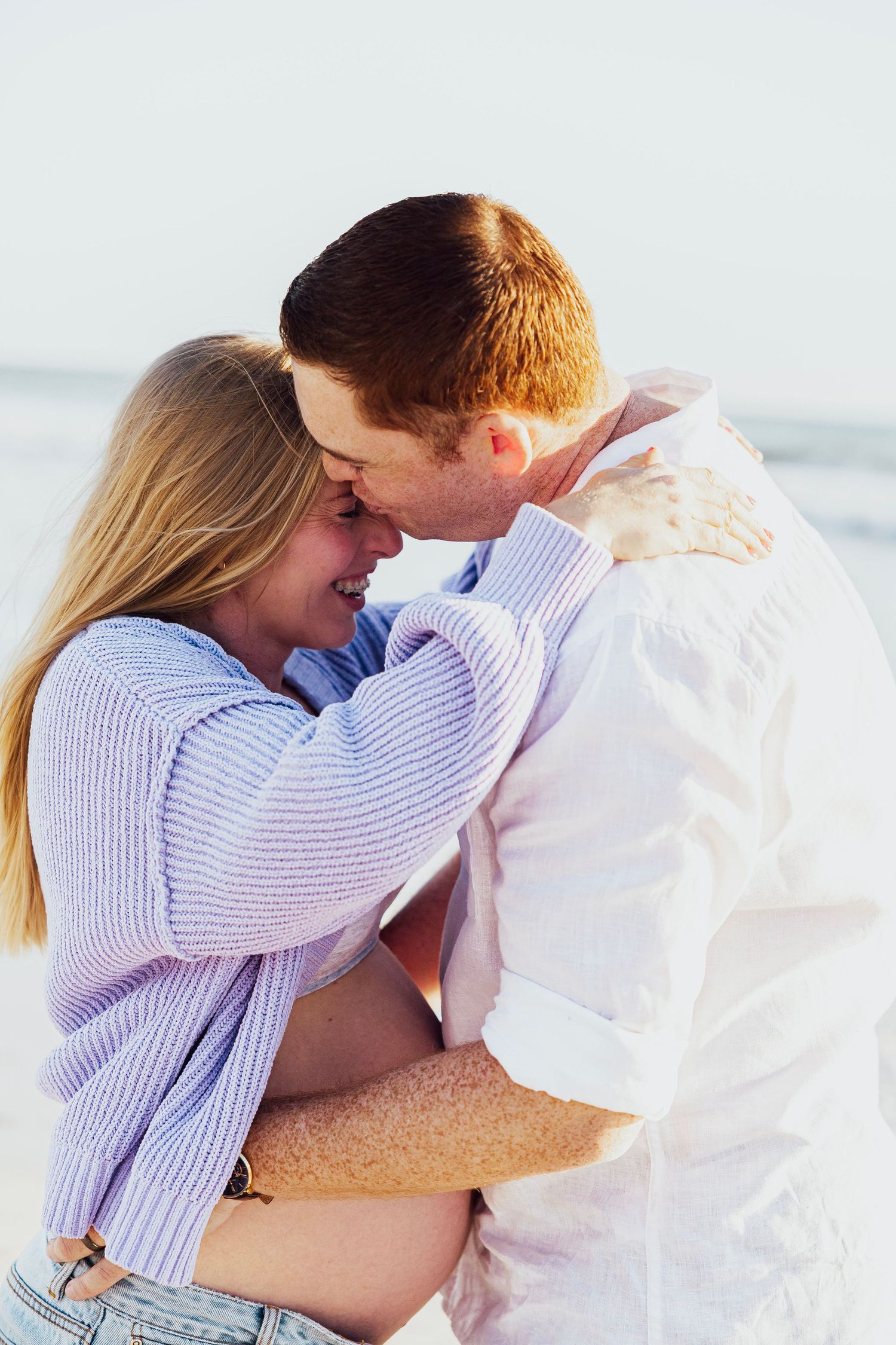 Pregnant couple embracing and touching foreheads on a bright sunny beach, woman in purple cardigan.