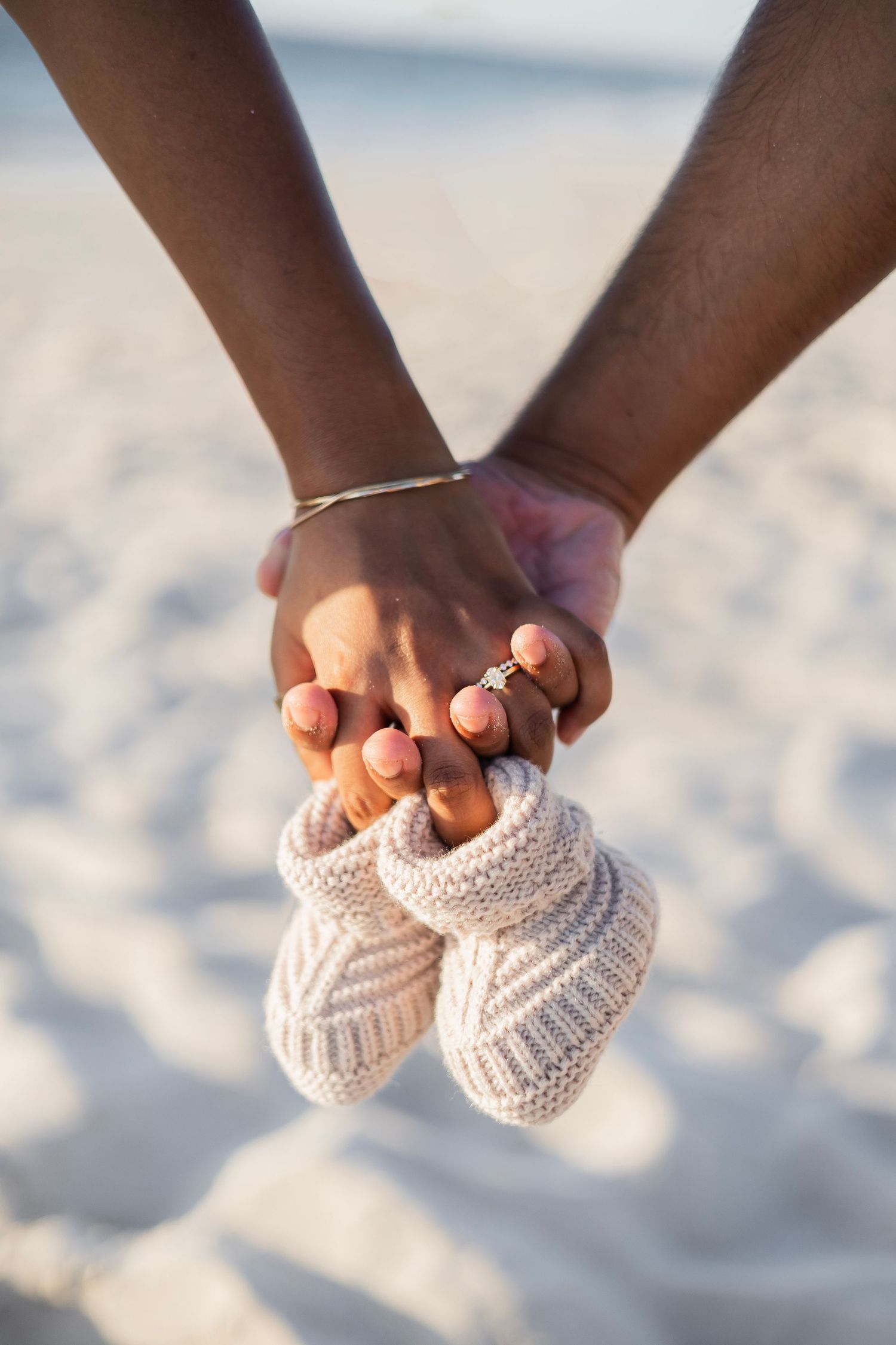 Couple holding hands together cradling tiny knitted baby booties on a sandy beach.