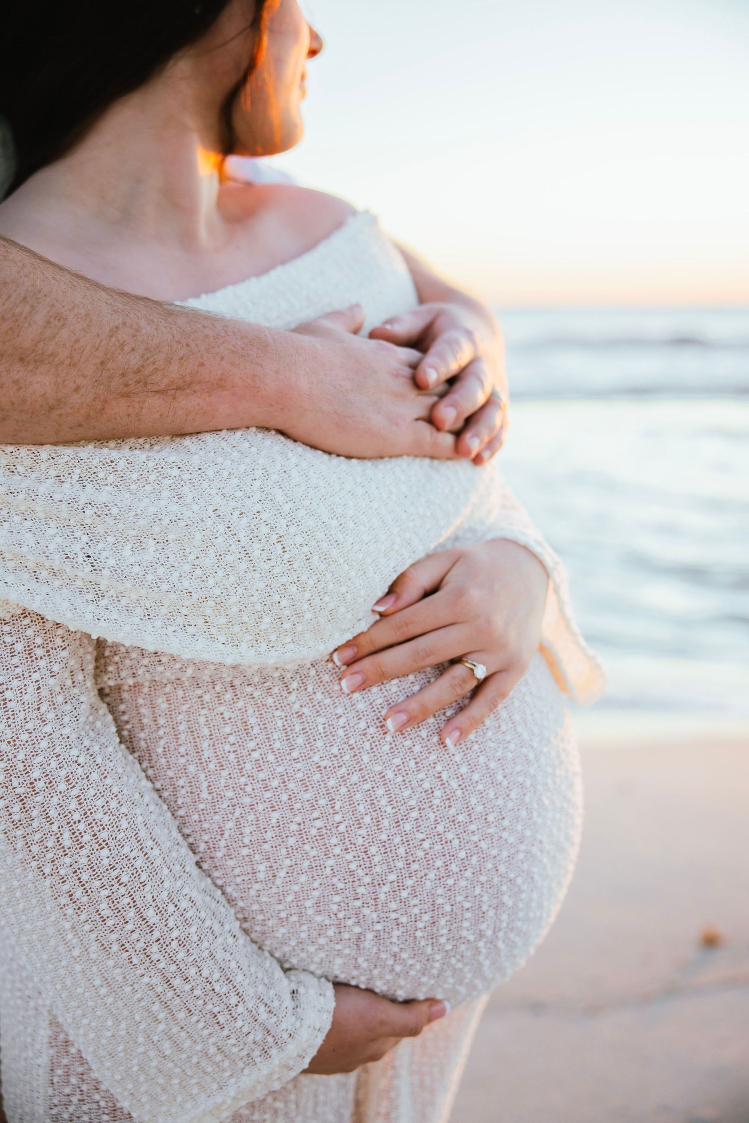 Side view of pregnant woman in white textured dress with partner's arms wrapped around her at sunset beach.