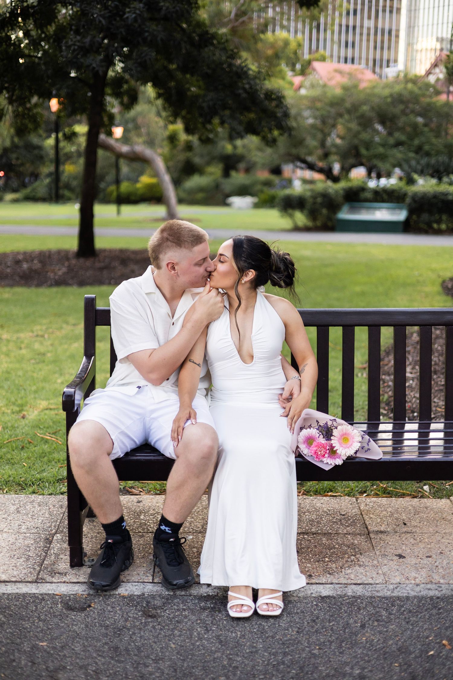 Couple kissing on park bench with bride holding pink flower bouquet.