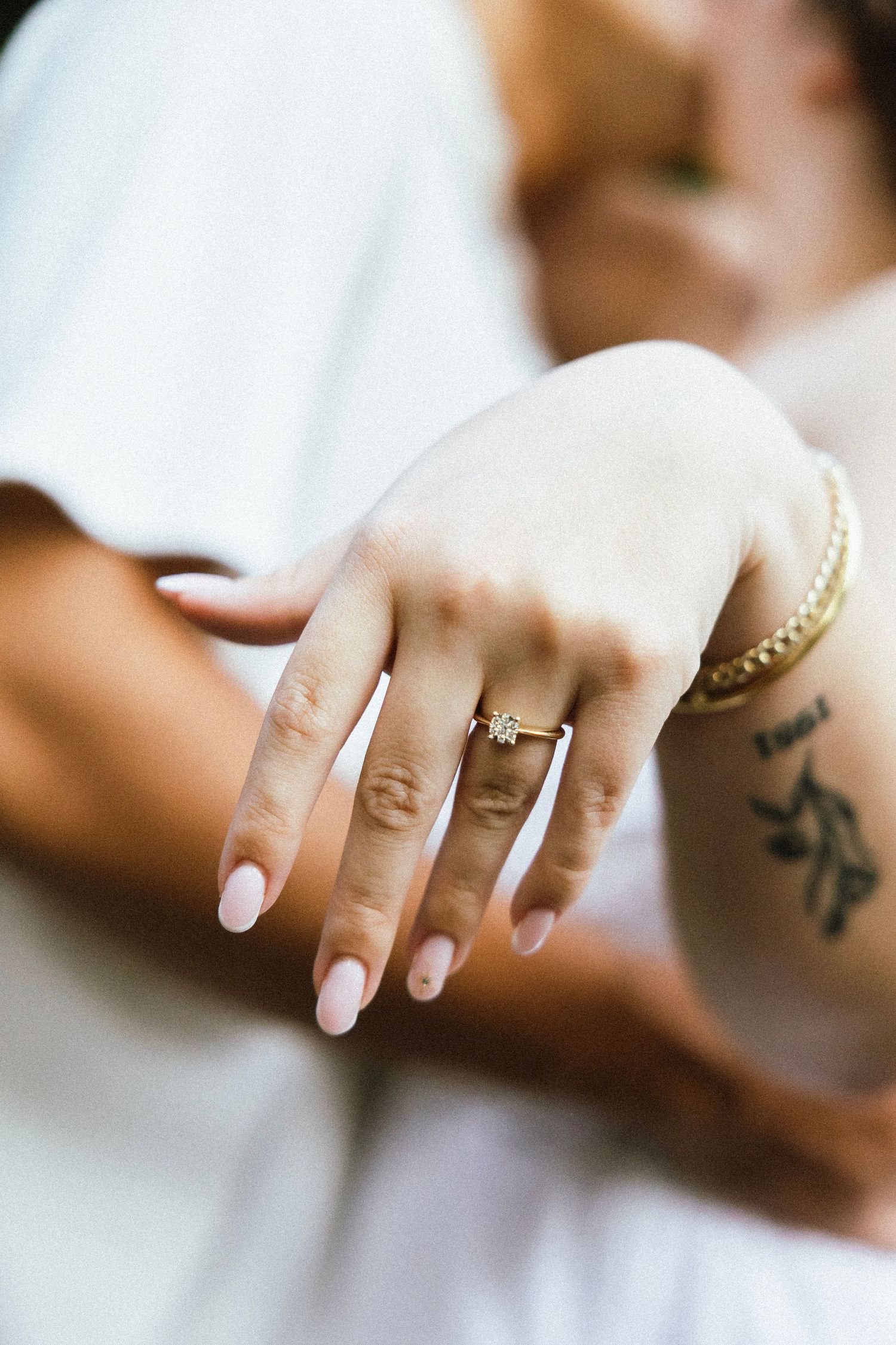 Close-up of an engagement ring on a womans hand with a gold bracelet and tattoo.