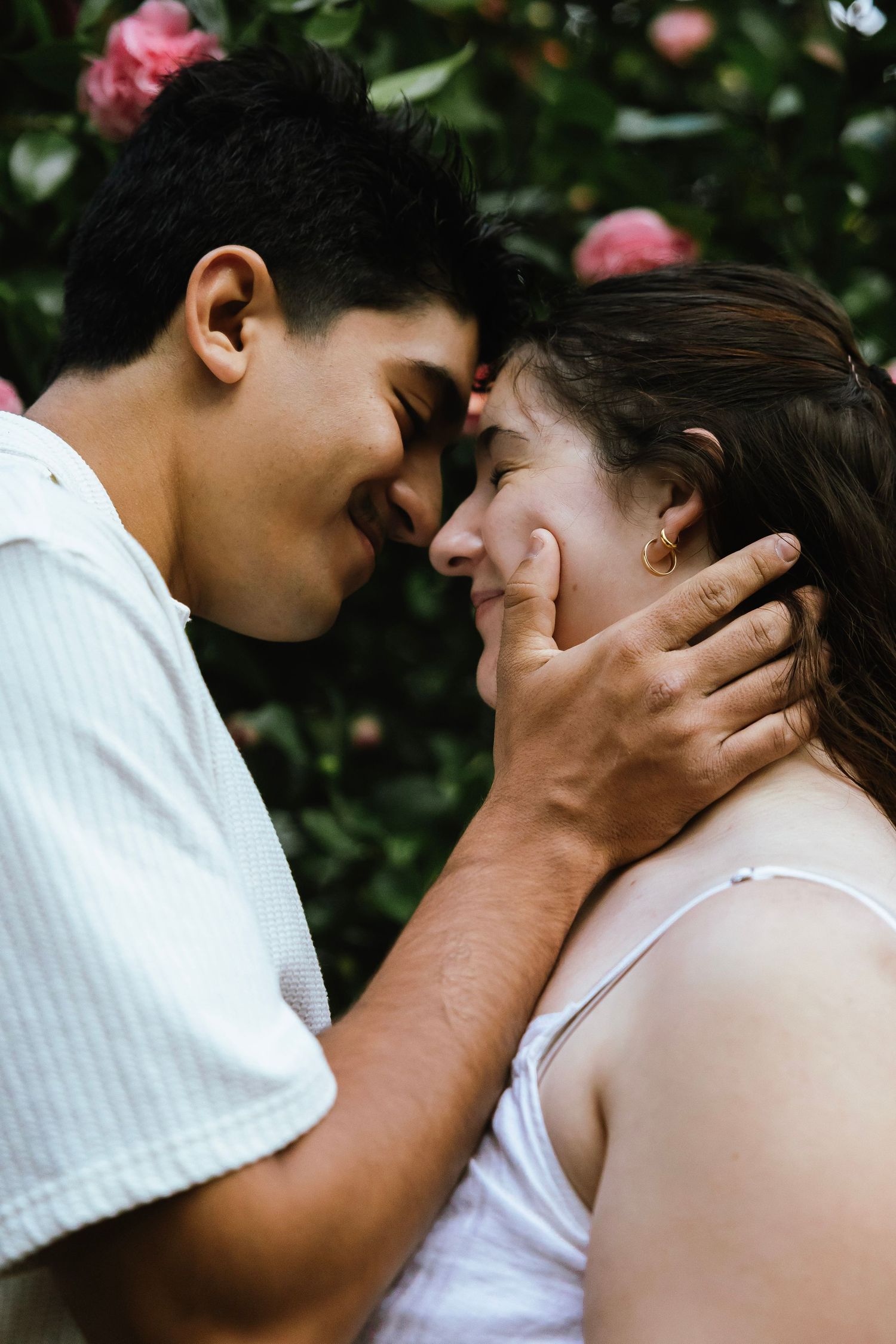 A couple shares a tender romantic moment outdoors, with the man gently cradling the woman's face near blooming pink roses.
