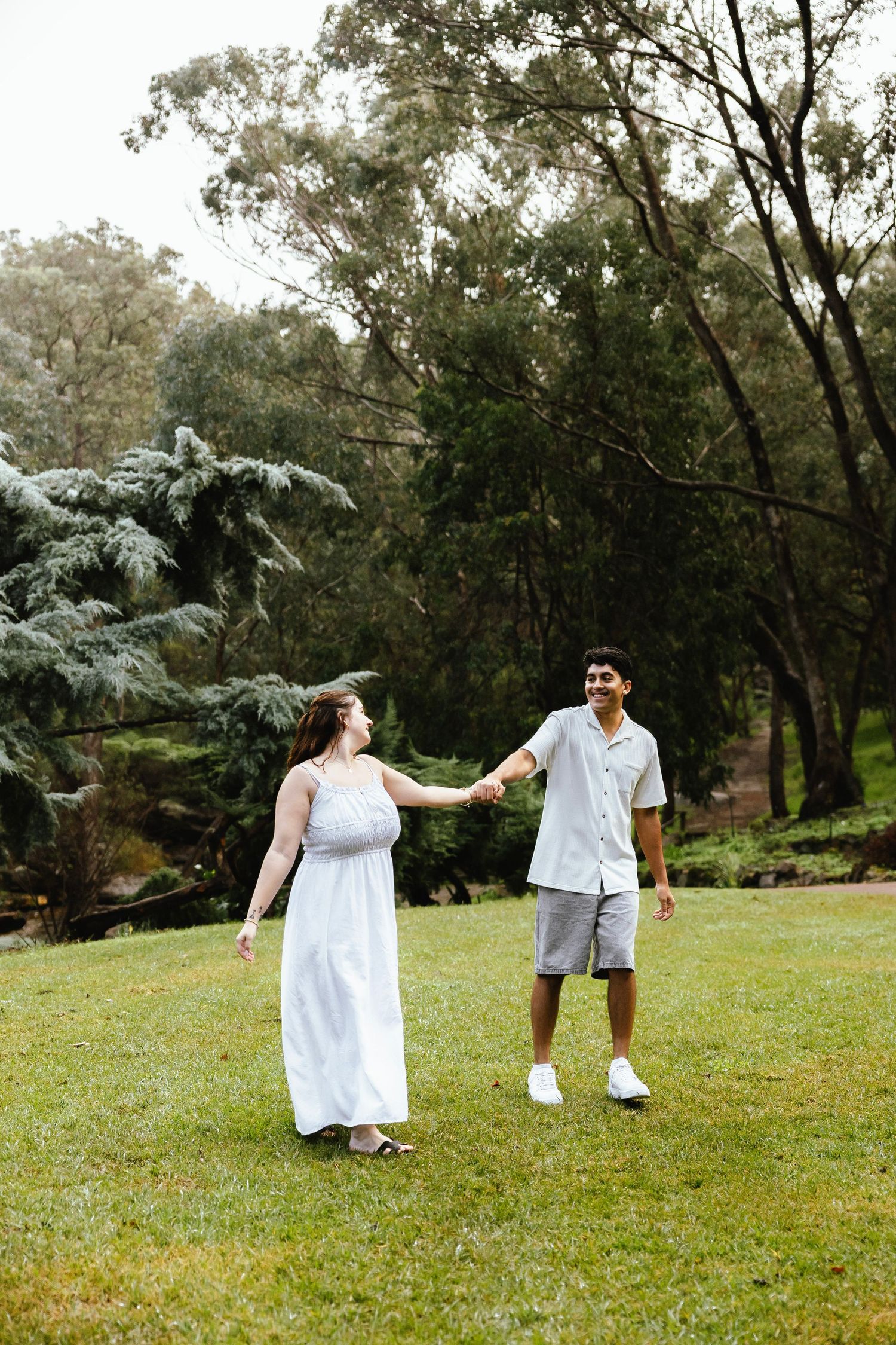 Couple holding hands and walking on green grass in a lush park, woman in white dress, man in casual shirt and shorts.