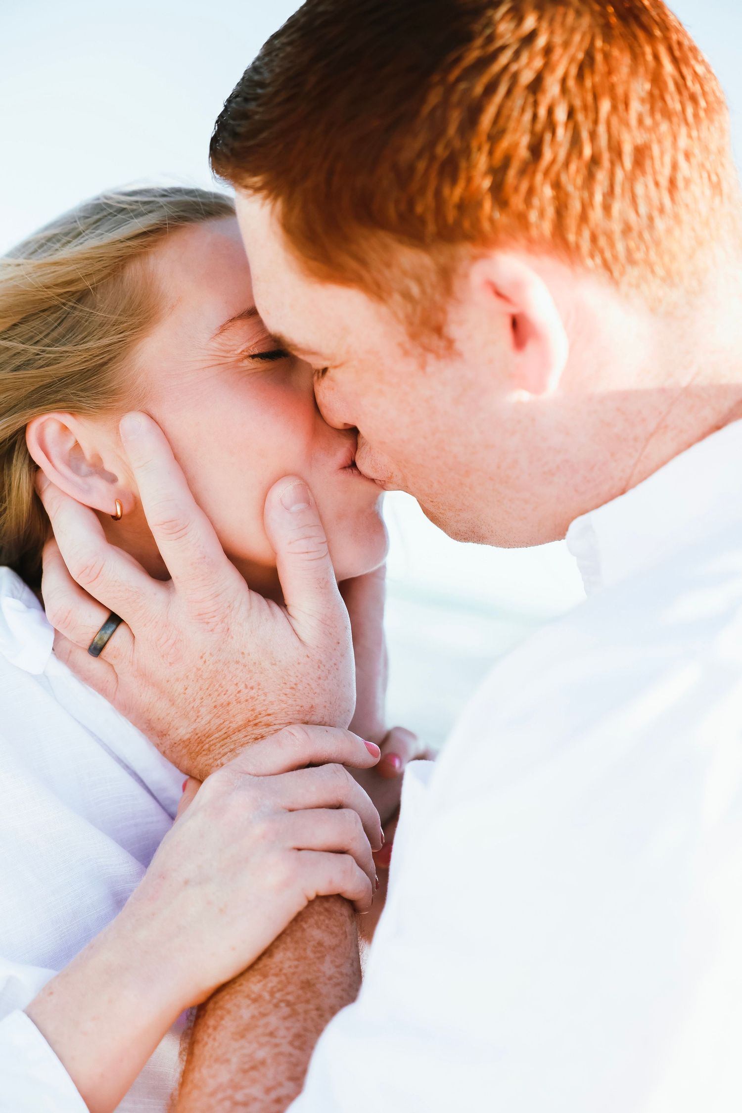 Close-up of a couple sharing a tender kiss on the beach, with an engagement ring visible on her hand.