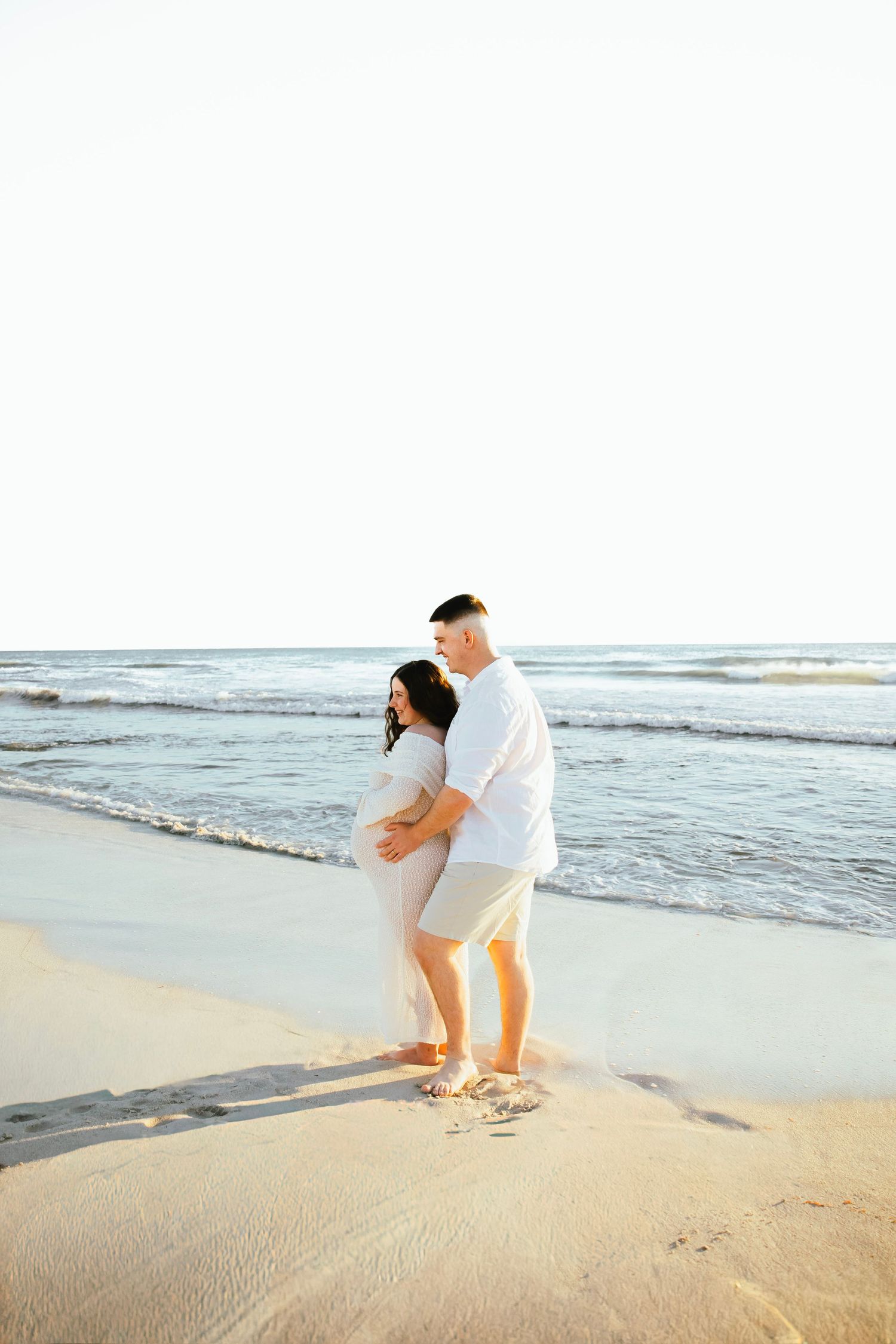 Pregnant couple embracing on sandy beach at sunset, man holding woman's baby bump near ocean waves.