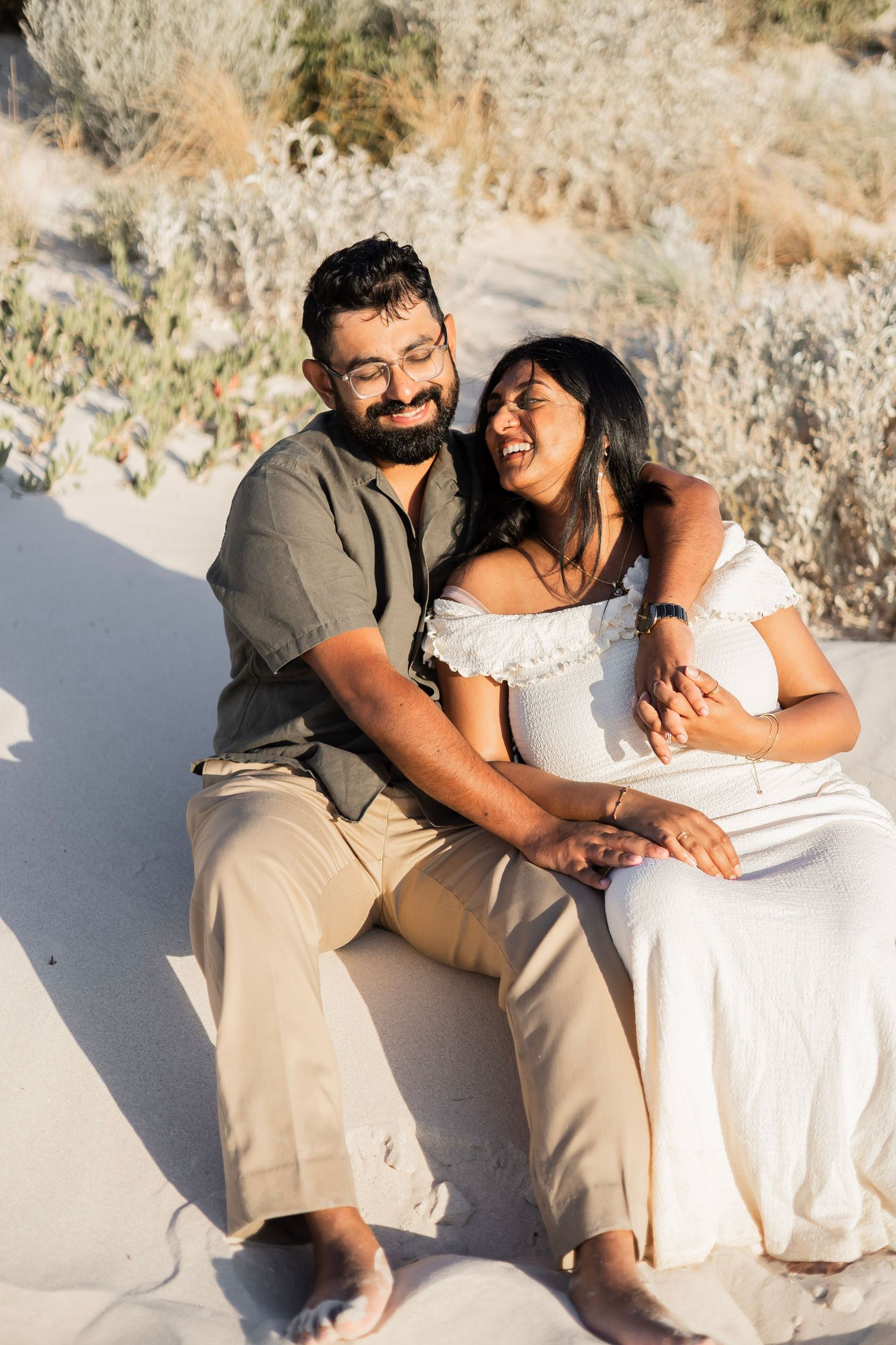 Happy couple laughing and cuddling together while sitting on sandy beach dunes at golden hour.