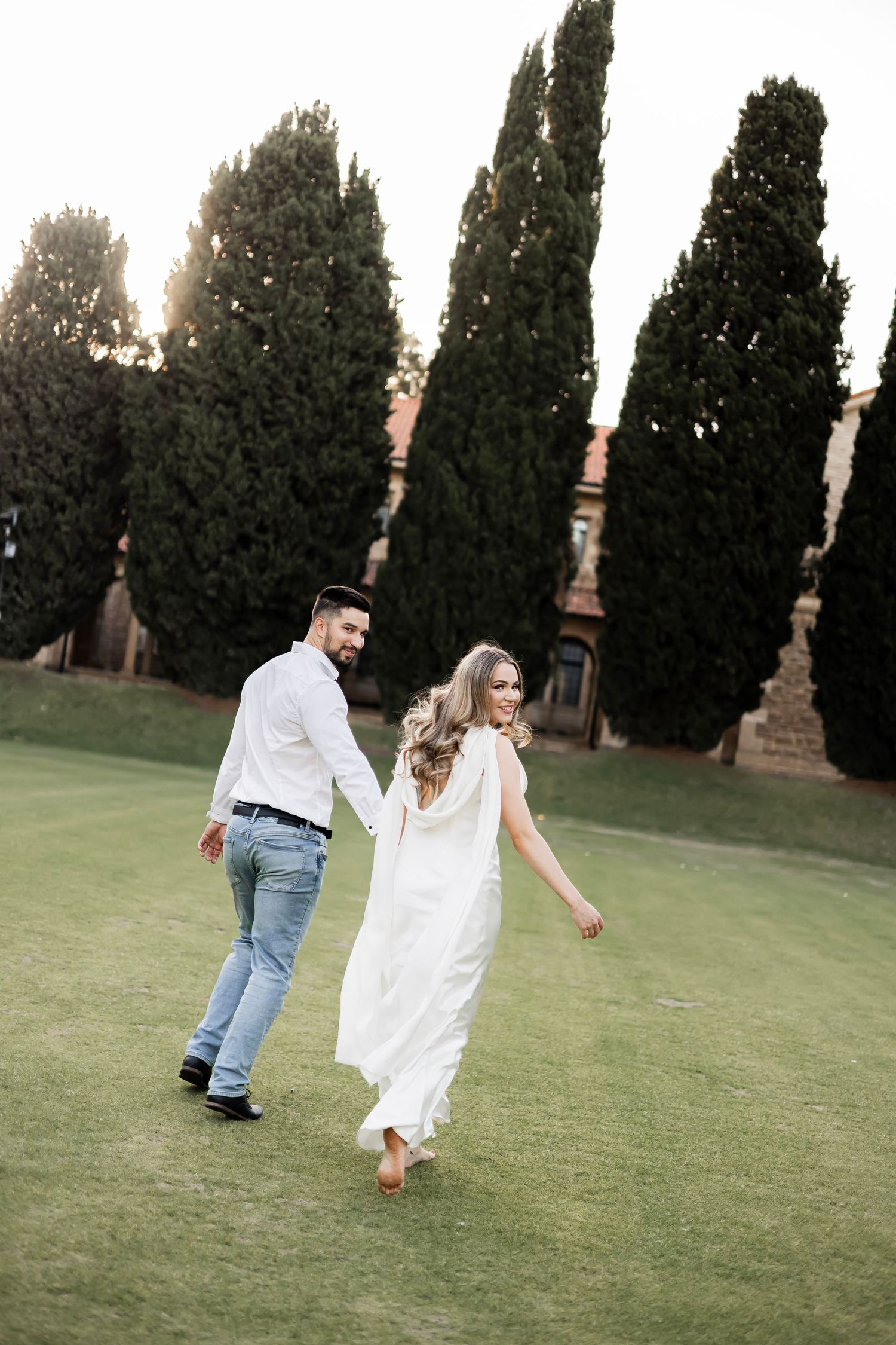 Couple walking hand in hand on green lawn with tall cypress trees, woman in white dress, man in jeans and white shirt.