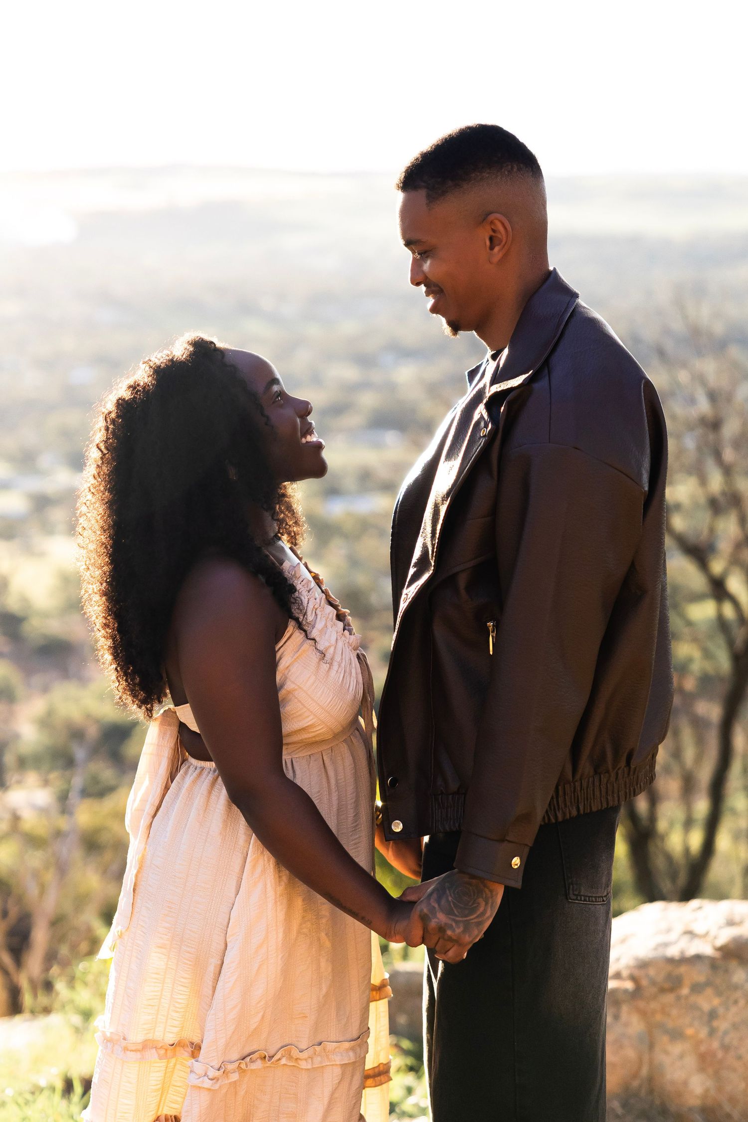 A couple holds hands and smiles at each other outdoors, with a scenic landscape and warm sunlight behind them.