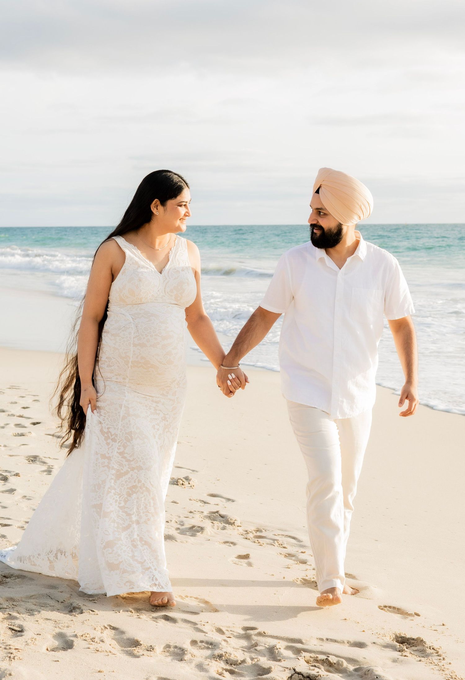 Smiling pregnant couple holding hands on beach, woman in white lace dress, man wearing a light pink turban.