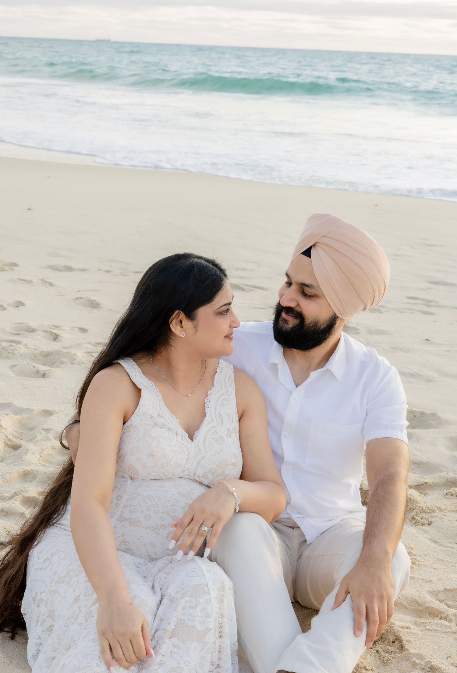 Expecting couple sitting on beach sand gazing lovingly at each other.