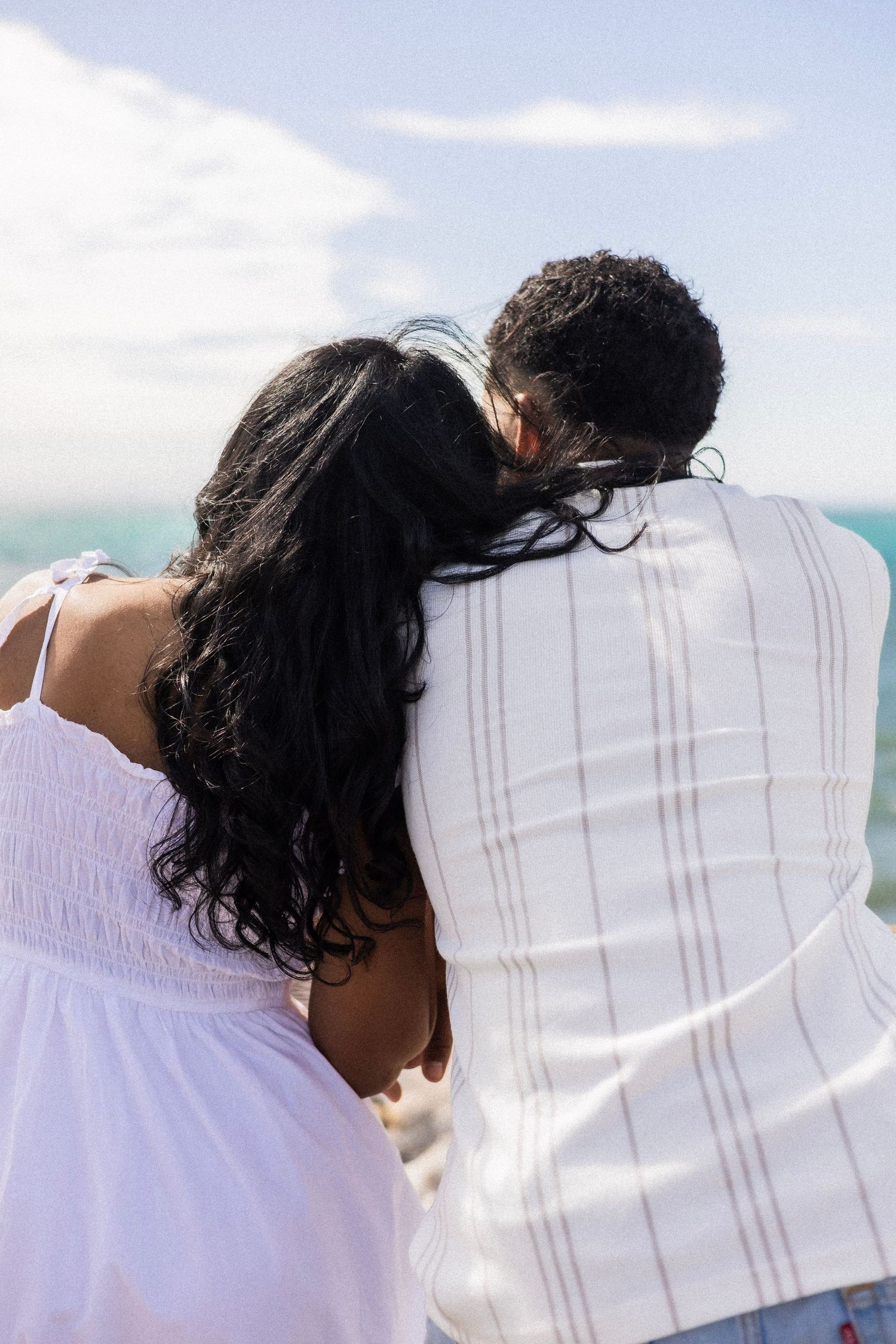 Back view of a couple in white embracing and leaning together while looking out at the ocean.