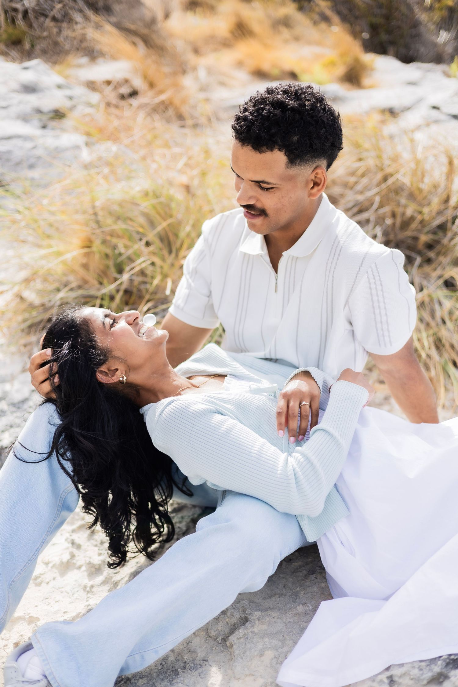 A laughing couple sits on beach rocks, the man tenderly holding the woman as she reclines.