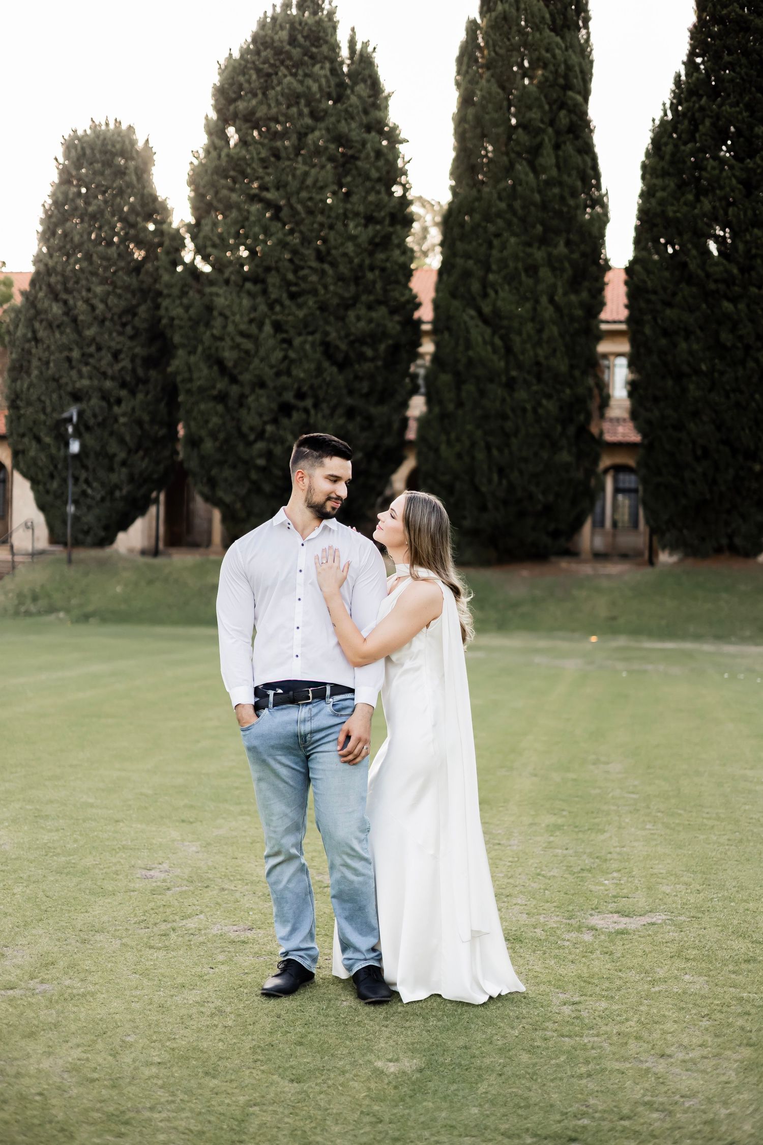 Couple sharing a tender moment on green lawn, woman in white bridal gown leaning toward smiling man in jeans.