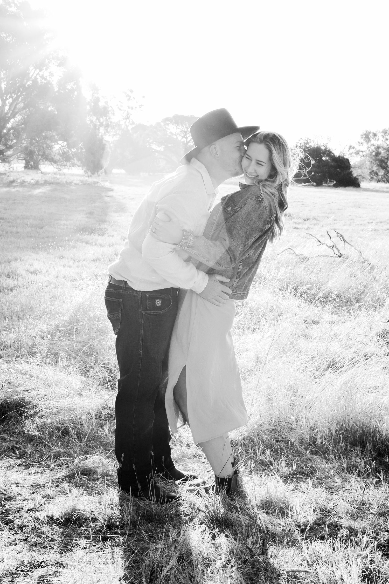 Black and white photo of a couple embracing and kissing in a sun-drenched open field with trees behind them.