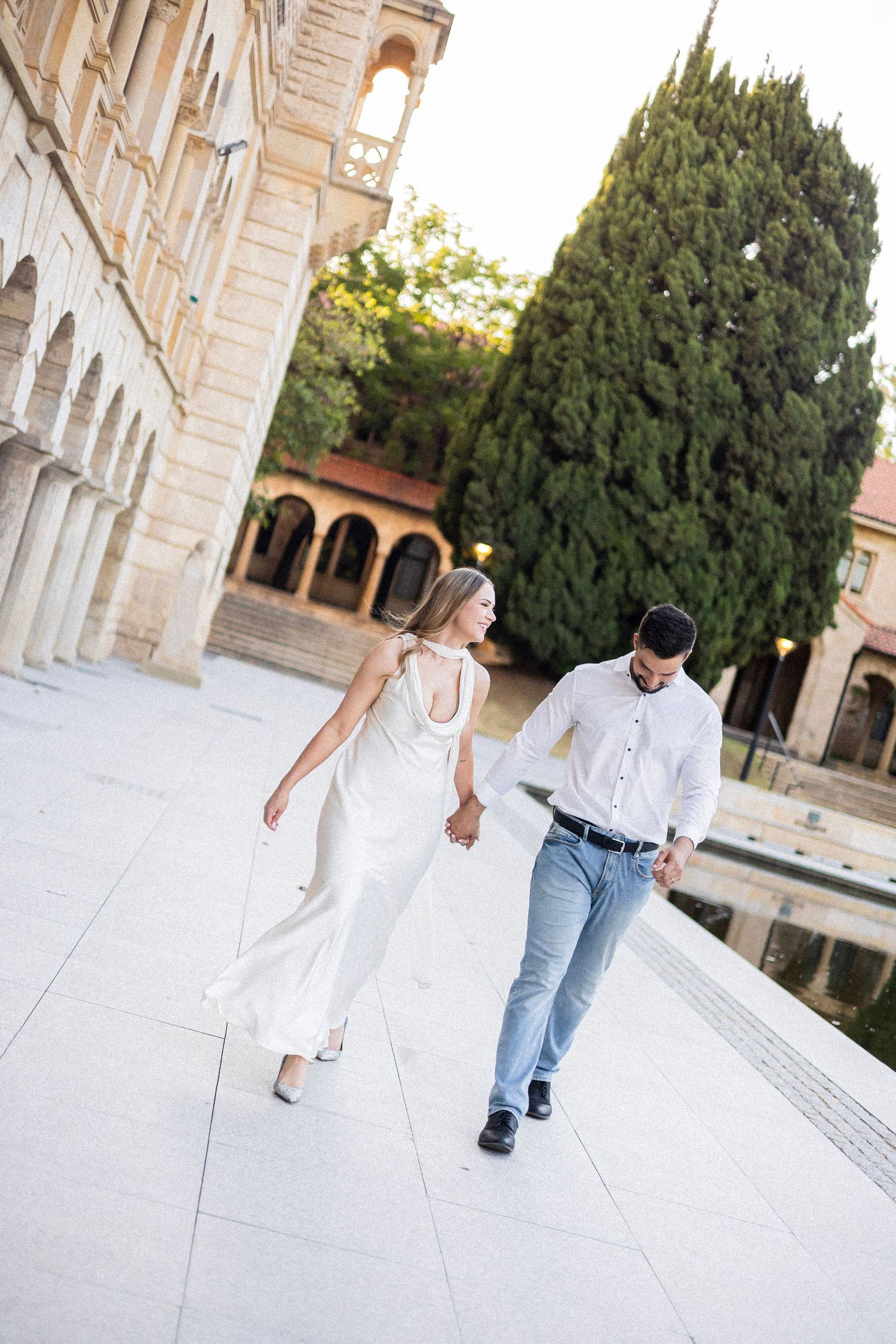 Couple in white attire walking hand in hand along elegant courtyard with arched building and cypress tree.