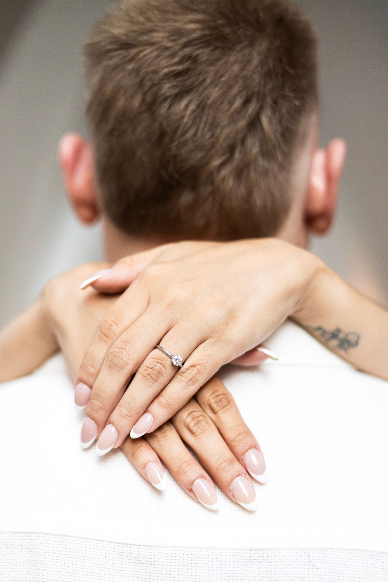Close-up of a womans hands with French manicure and diamond engagement ring resting on a mans shoulders.