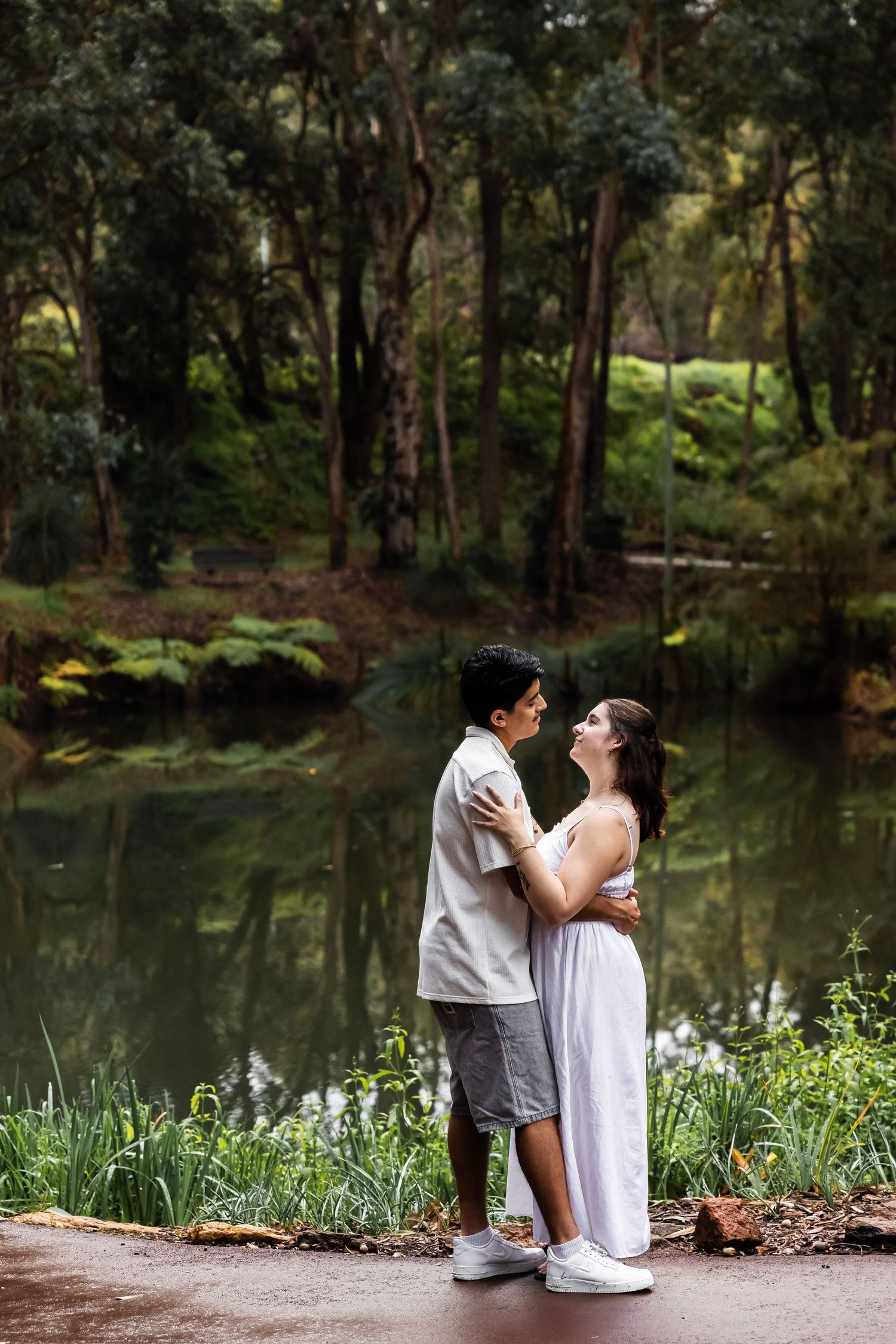 Couple embracing romantically by a serene forest pond, surrounded by lush green trees and ferns.