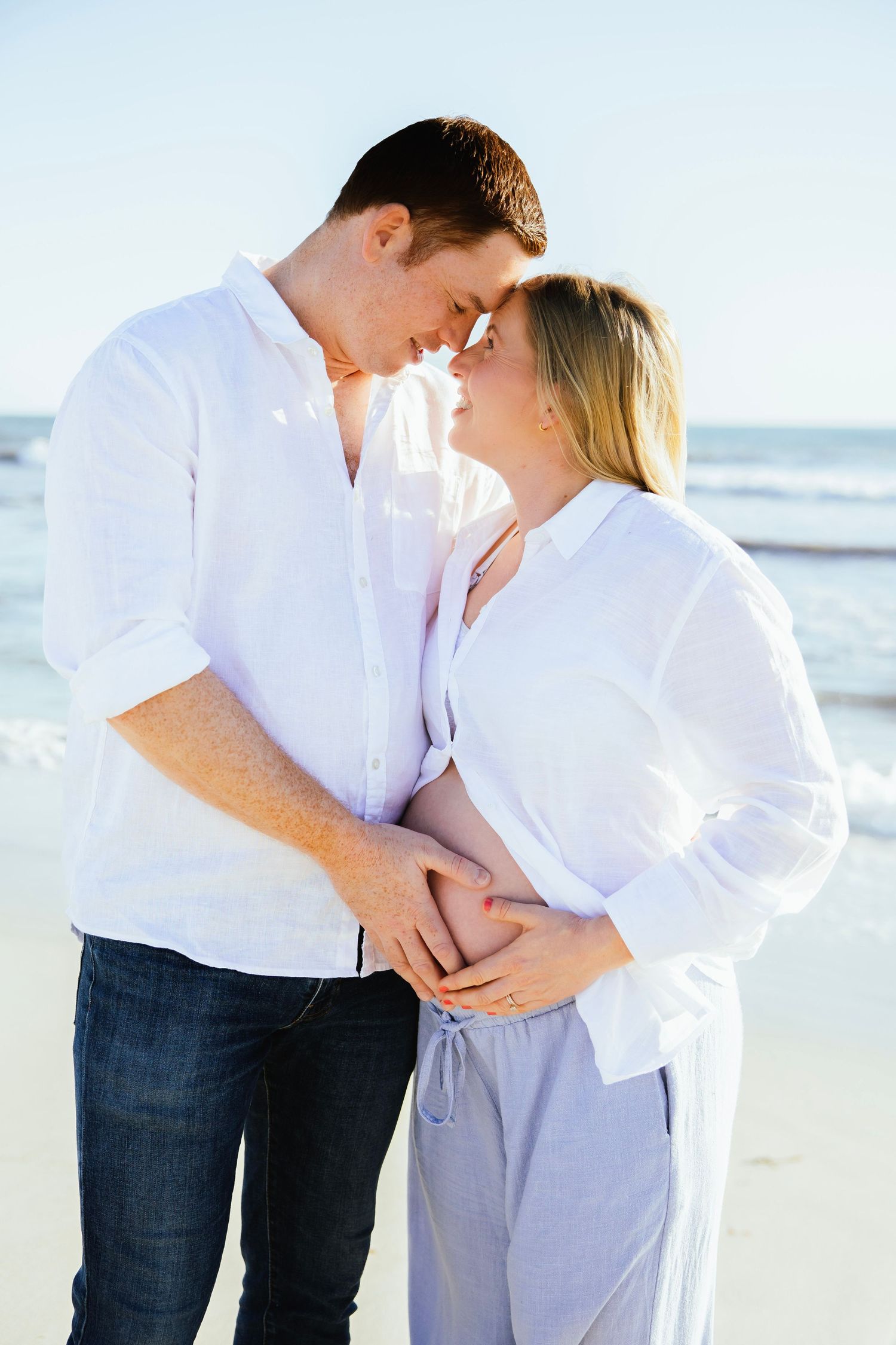 Couple touching foreheads tenderly on beach, both cradling pregnant belly together.