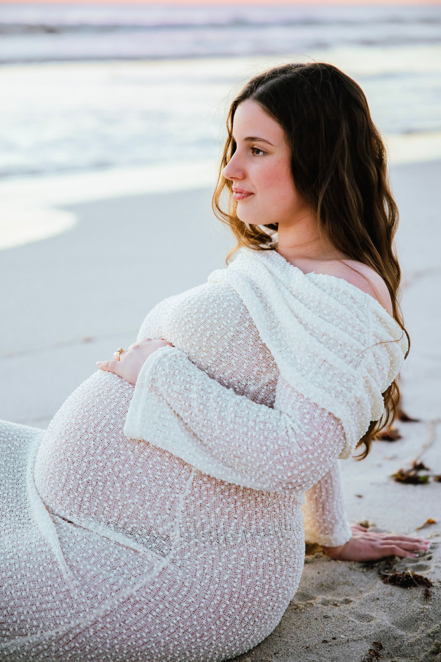 A pregnant woman in a sheer white gown sits on a sandy beach at sunset.