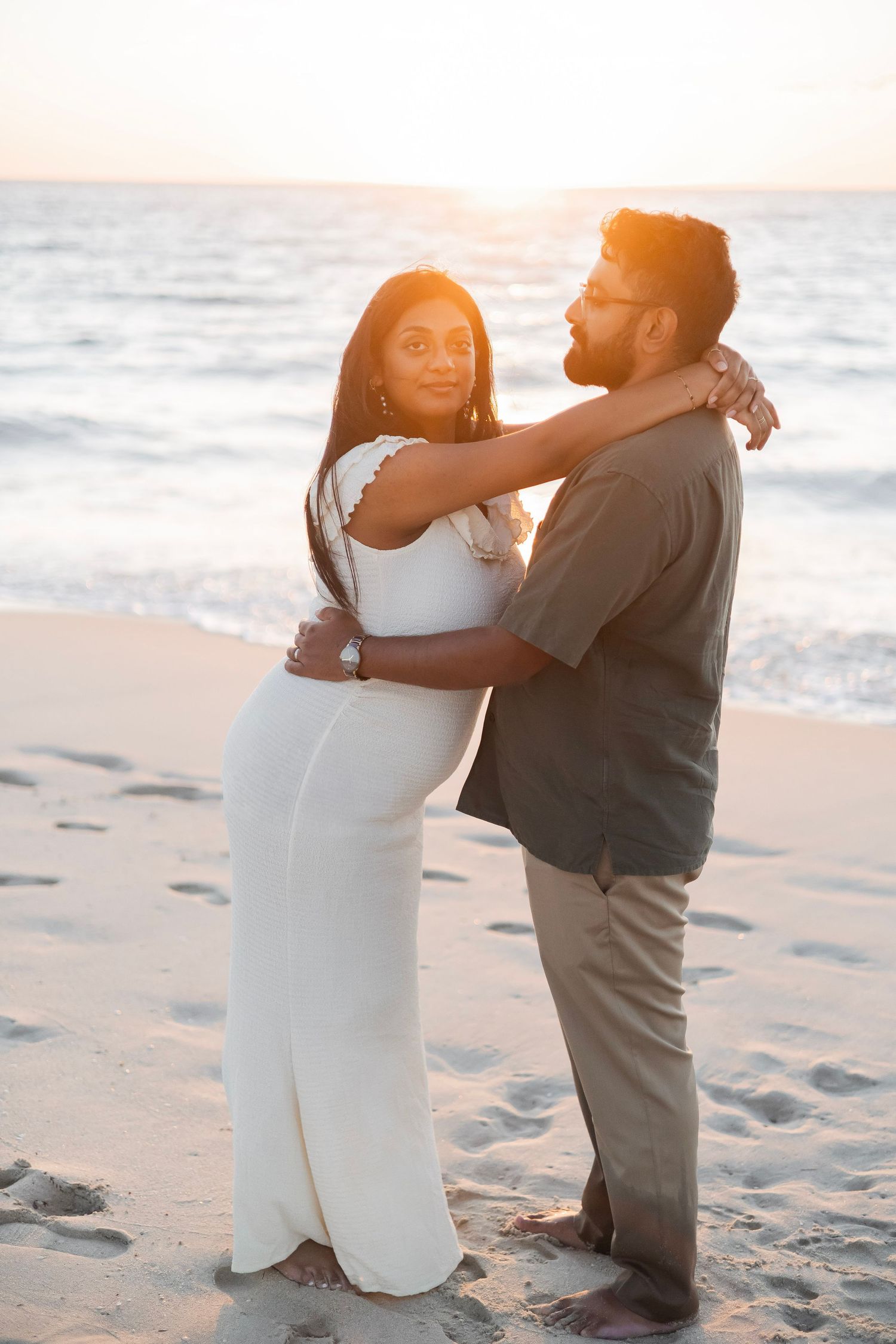 Couple embracing on sandy beach at golden sunset, woman in white dress with arms around man in gray shirt.
