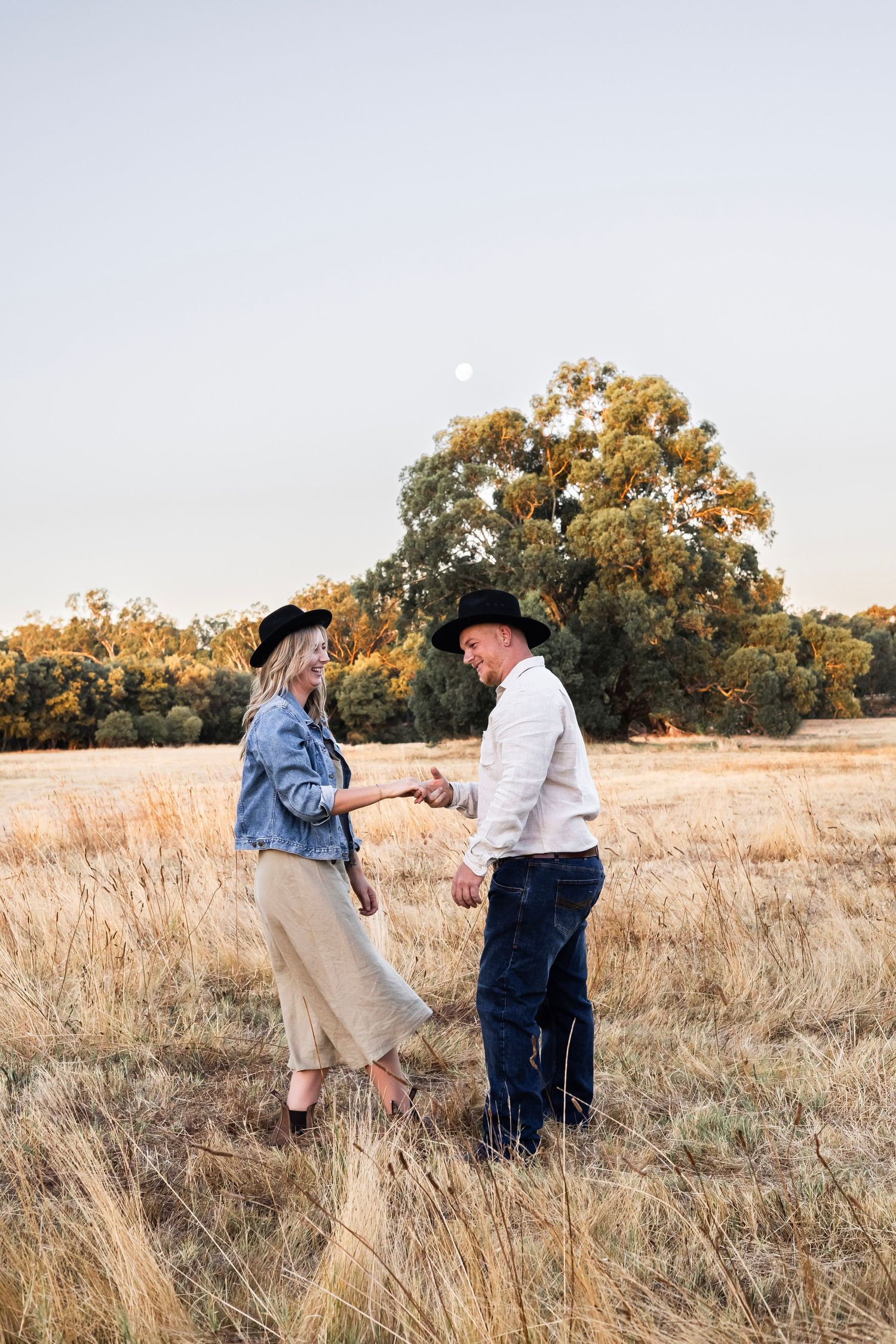 Couple holding hands and dancing together in a golden field at dusk with a full moon.
