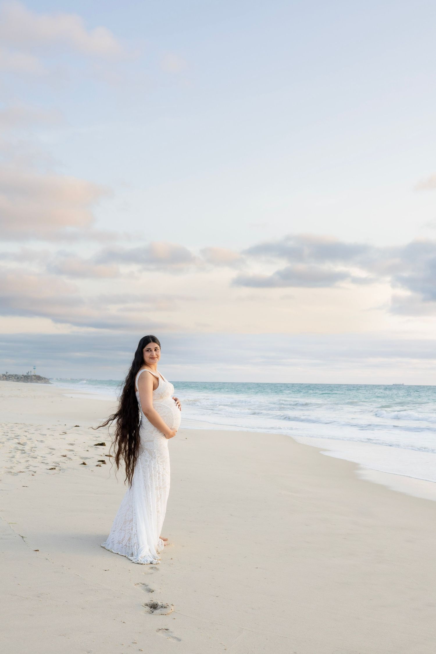 Pregnant woman in white lace dress cradling baby bump on sandy beach at sunset.