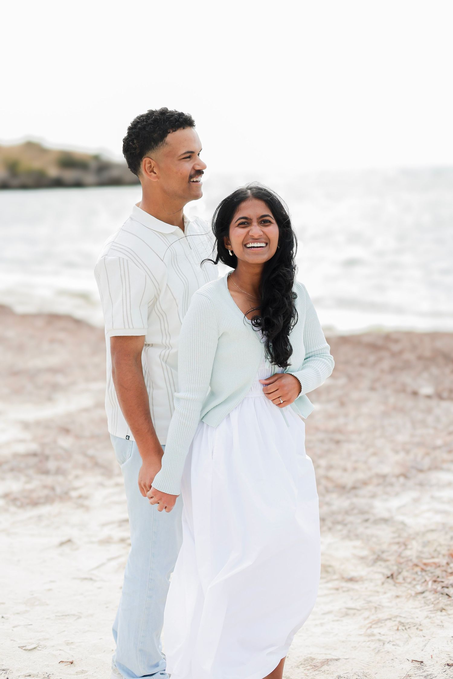 Smiling couple standing close together on a beach with calm ocean water in background.