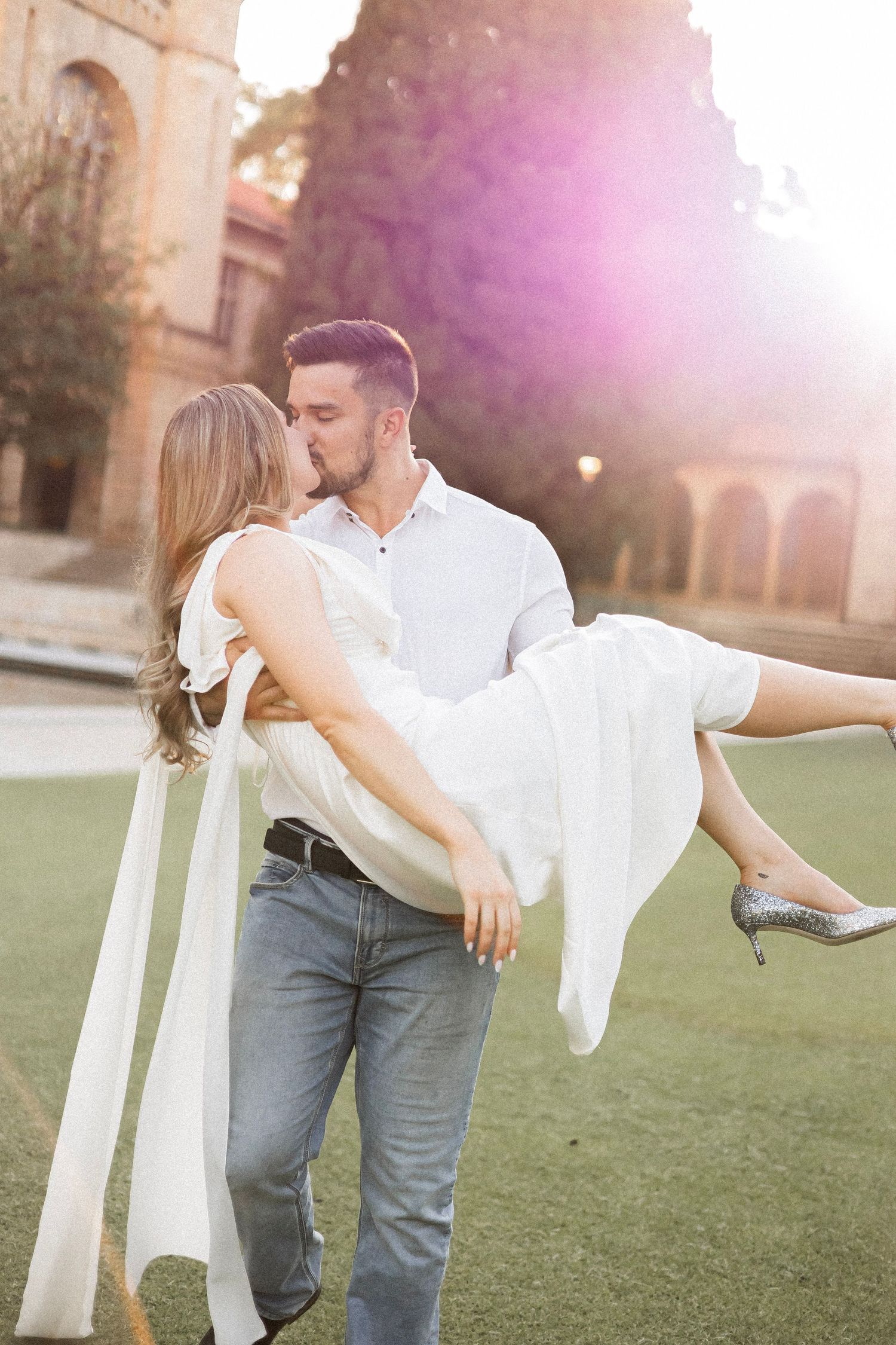 Couple sharing a romantic kiss as groom carries bride in white dress outdoors in golden sunlight.