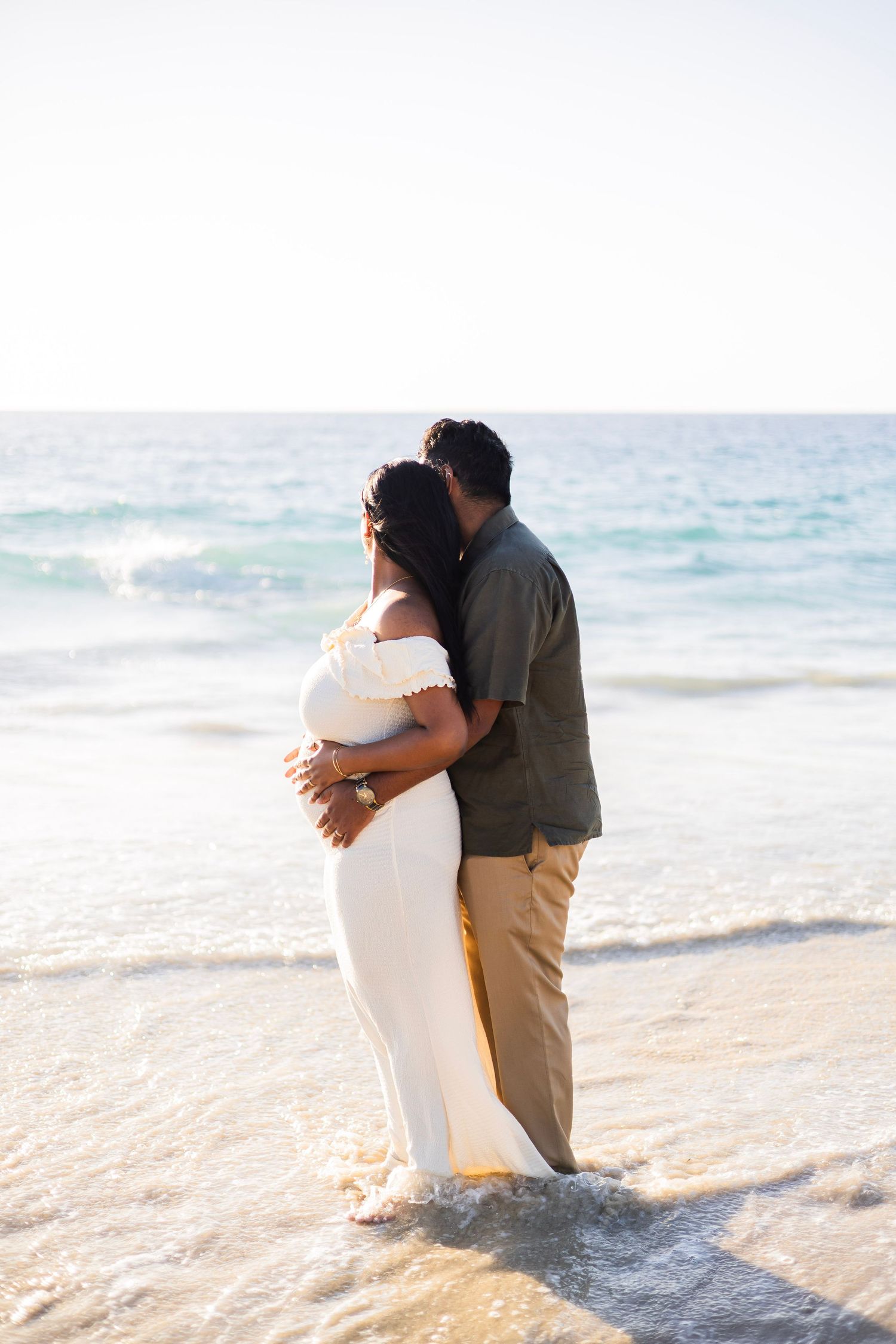 Couple embracing on a sunny beach, woman in white off-shoulder maternity gown, ocean waves behind.