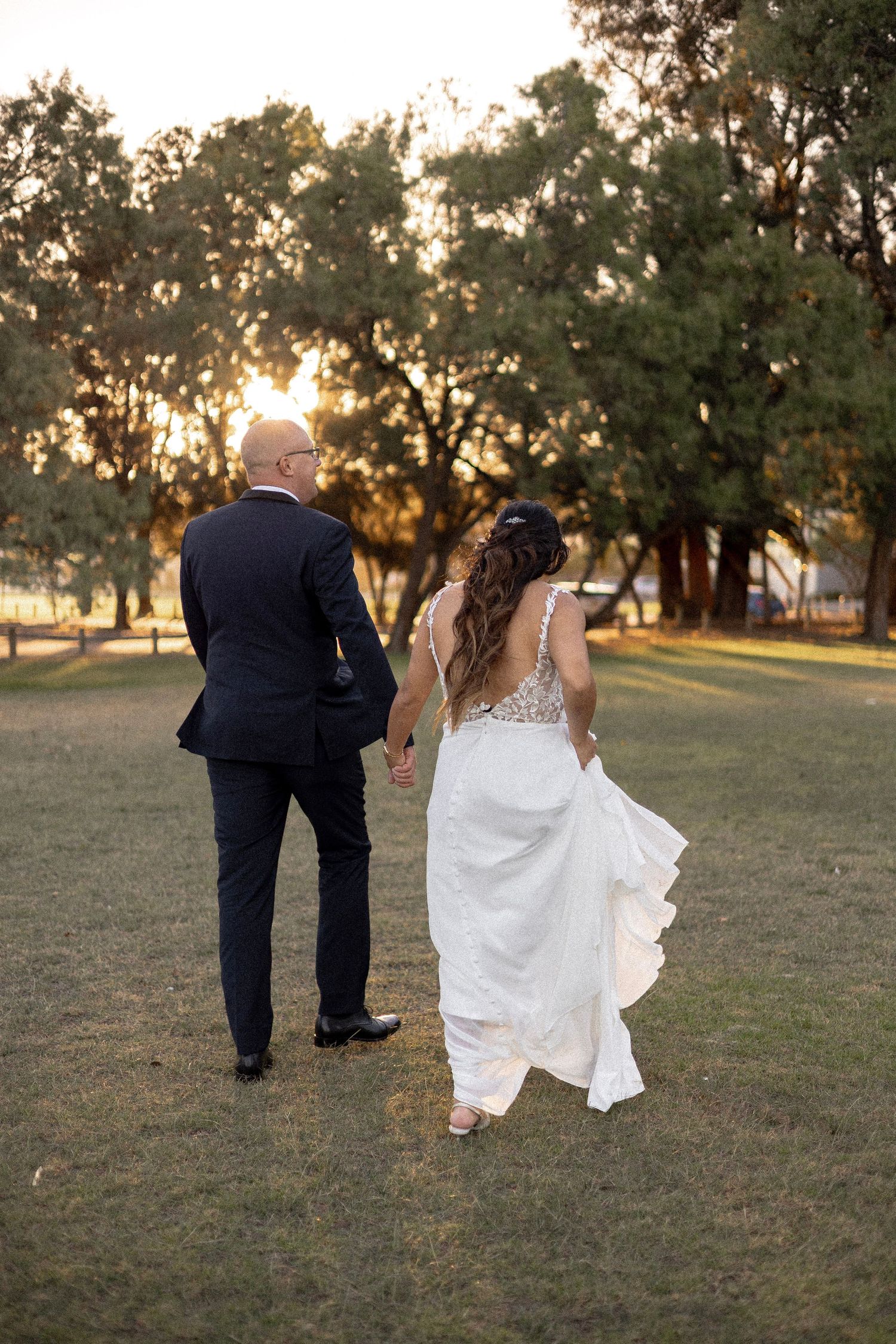 Bride and groom walk hand in hand toward golden sunset through tree-lined park after wedding ceremony.